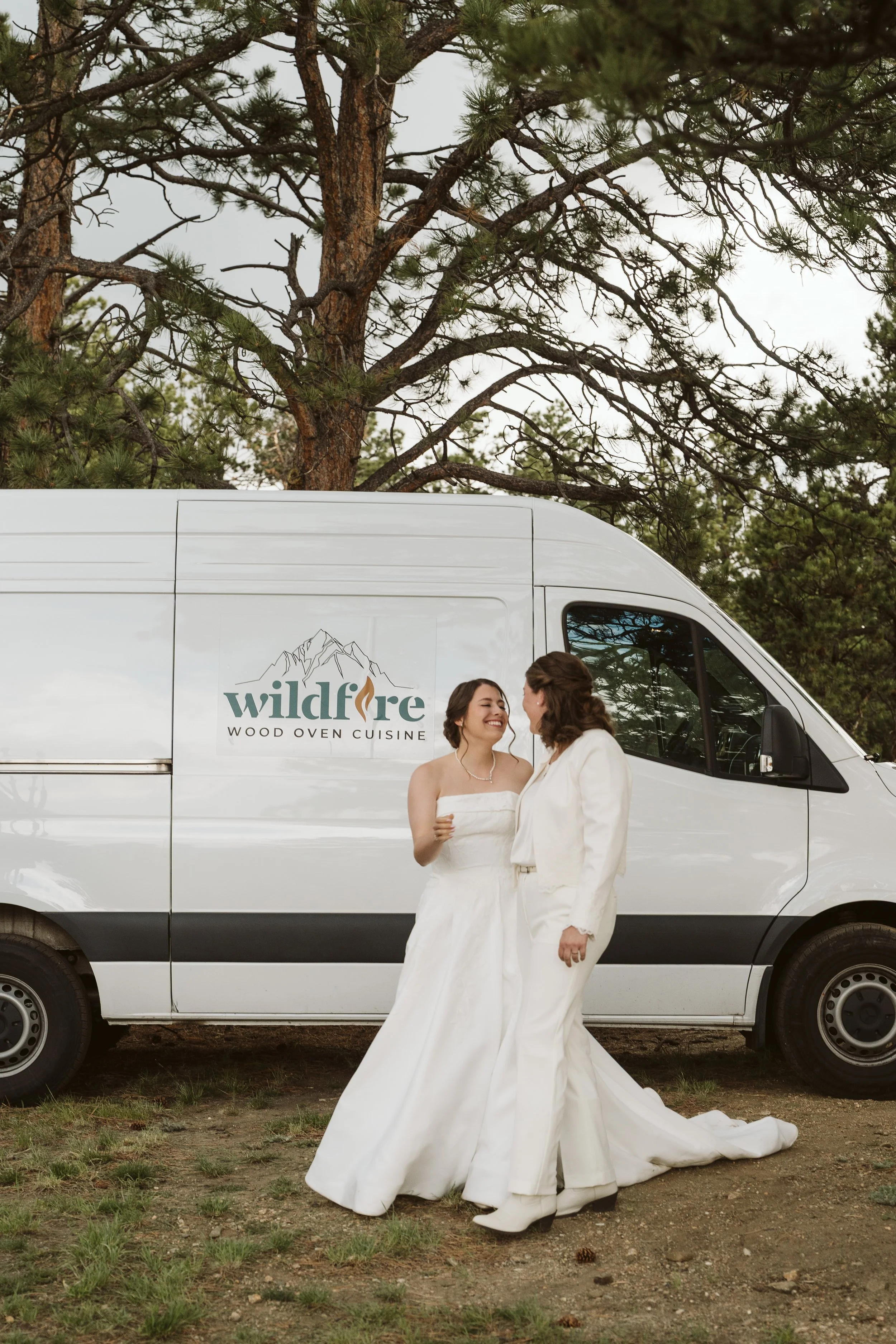 Two women in wedding dresses standing in front of a white van with a tree in the background. Guests enjoying Wildfire Cuisine Co. wood-fired catering at a Colorado wedding