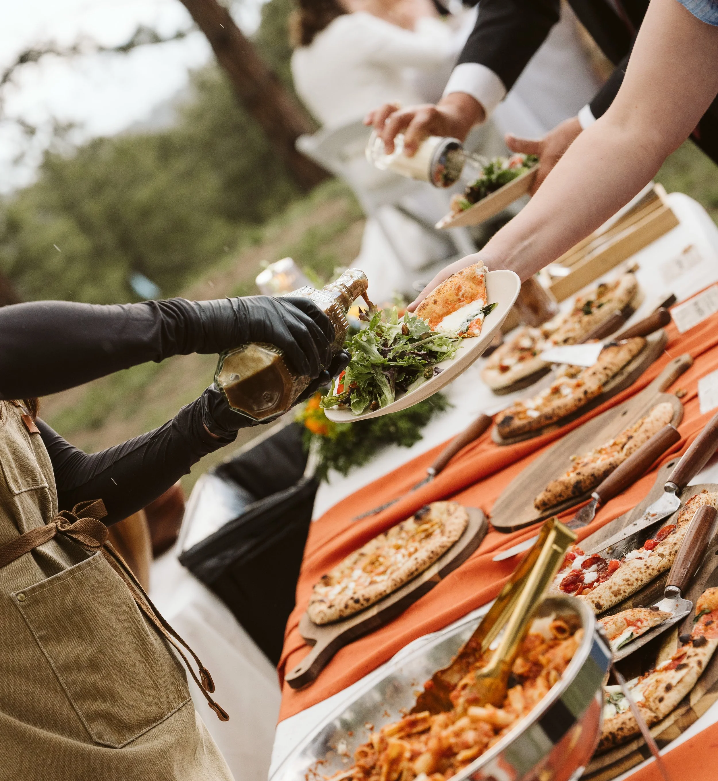 People serving themselves pizza from a buffet table outdoors.