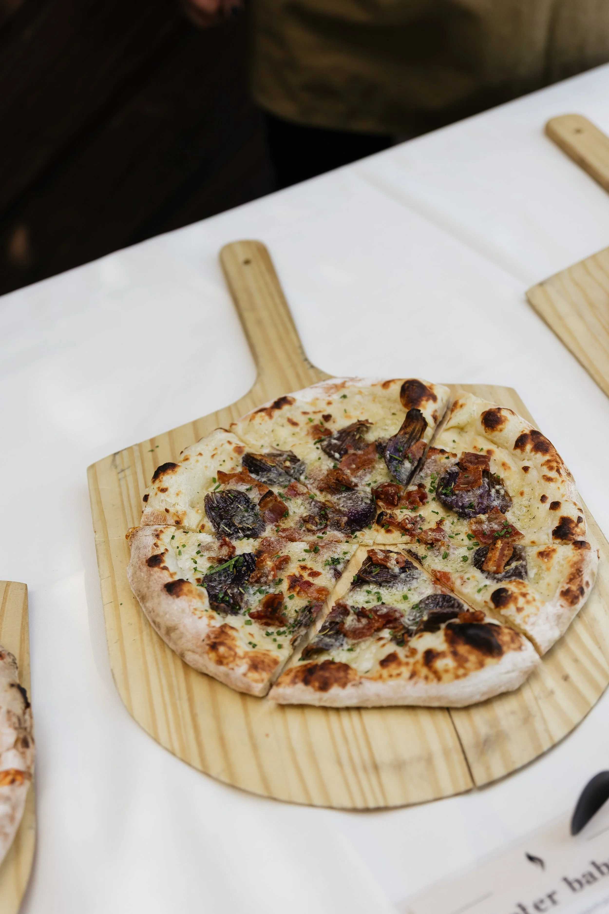 Close-up of a pizza with mushrooms, bacon, and herbs on a wooden pizza peel. Wood-fired wedding catering buffet in Denver by Wildfire Cuisine Co.