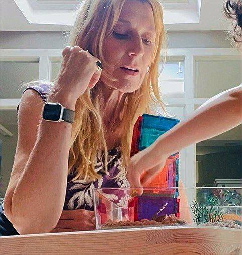 A woman with blonde hair looking focused while observing a child's hand playing with colorful building blocks inside a clear container.