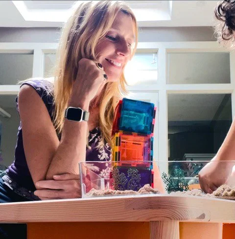 Woman with blonde hair smiling while playing with colorful translucent blocks on a table.