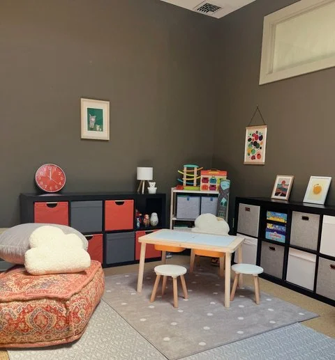 Children's playroom with a gray wall, colorful storage cubes, a small table with stools, and various toys and pictures.