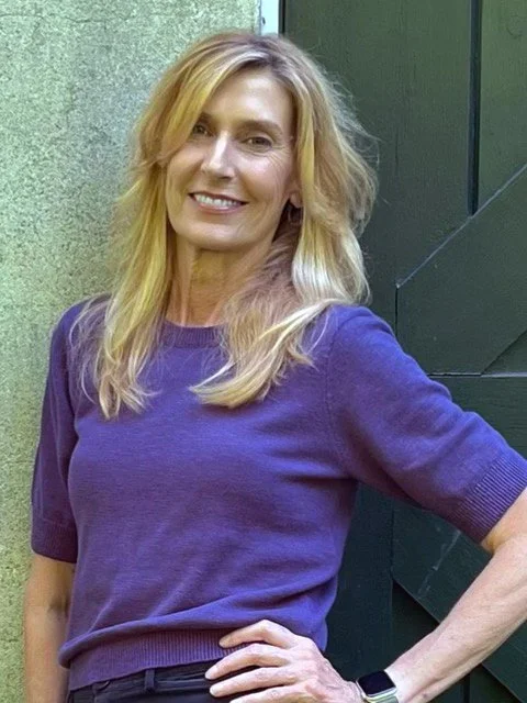 A woman with blonde hair, wearing a purple top and black pants, stands outdoors with a hand on her hip, smiling.