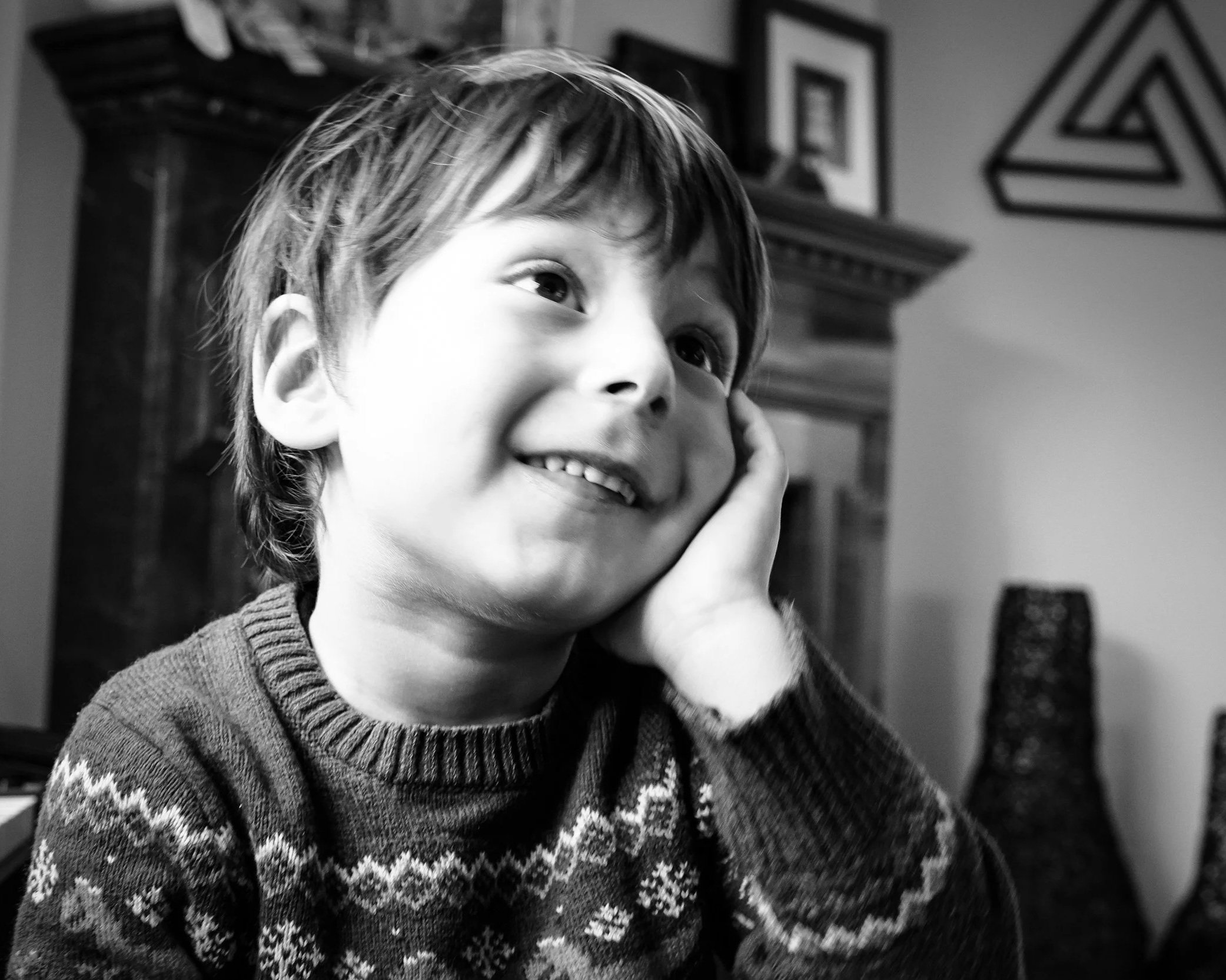A black and white photo of a young boy with short hair, smiling and resting his face in his hand, wearing a festive sweater with snowflake patterns.