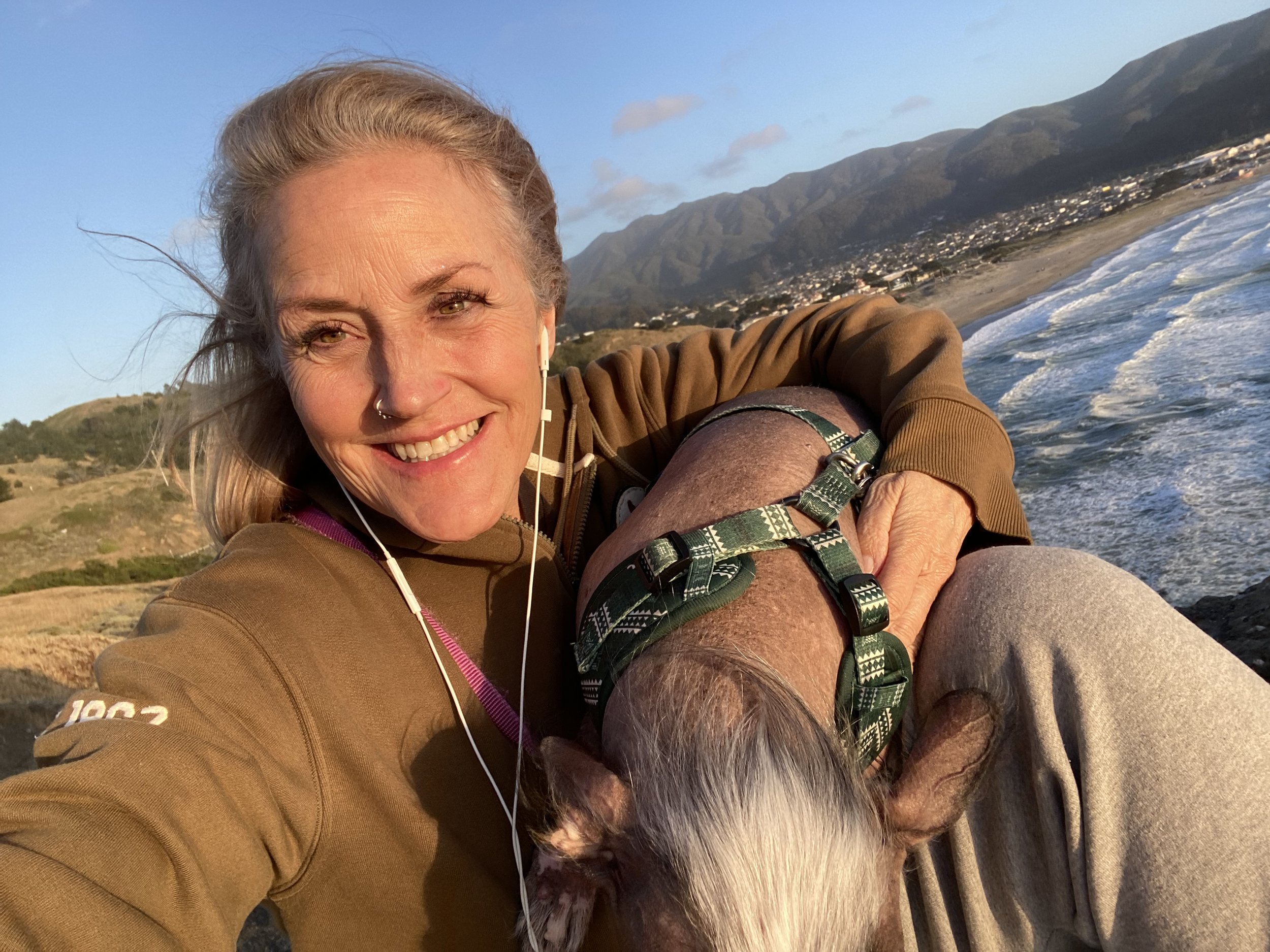 Woman smiling outdoors on a beach at sunset with mountains and dog beside her