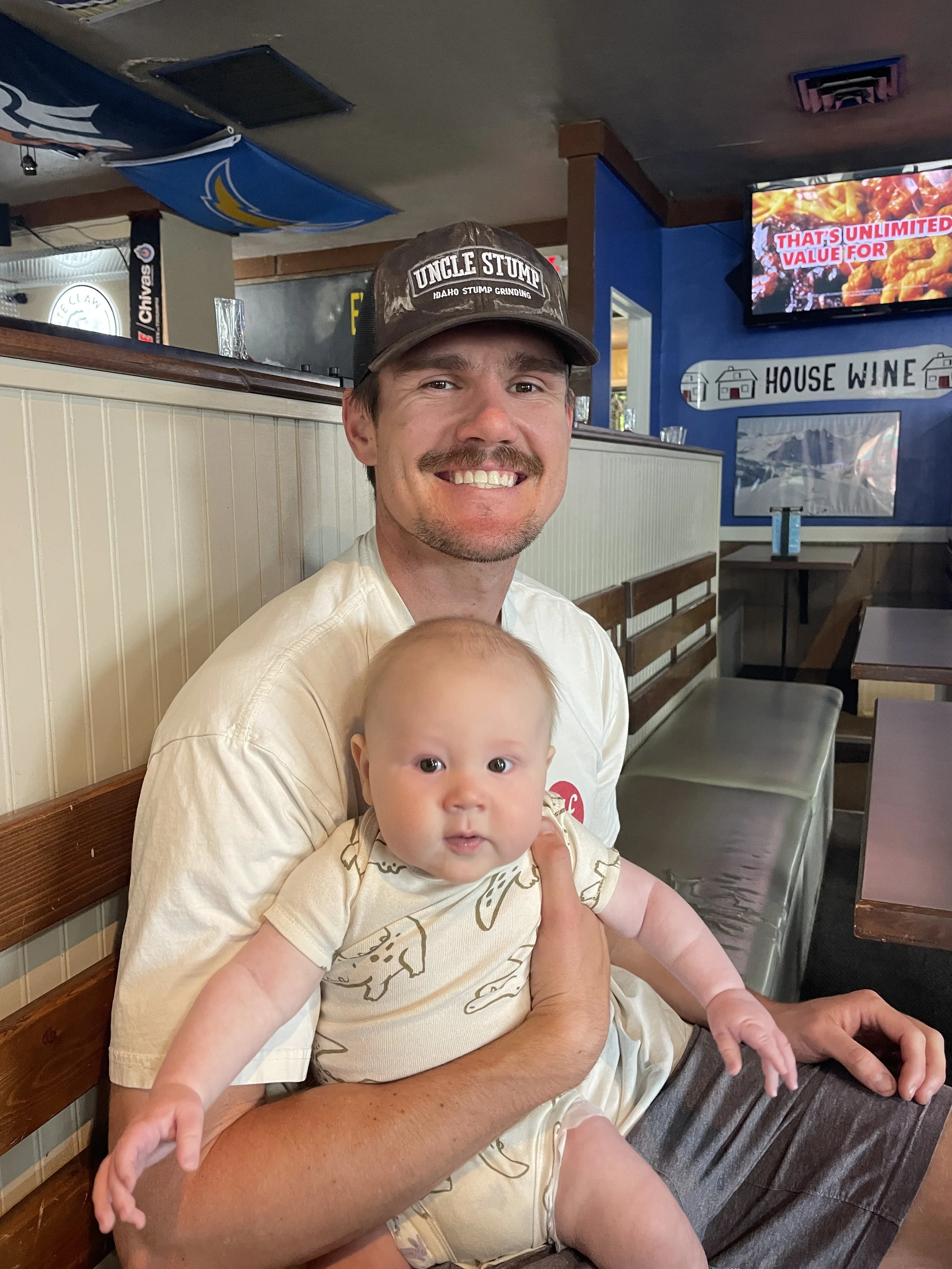 A man with a mustache and a baseball cap smiling while holding a baby with big eyes, seated in a restaurant with blue walls and a TV screen in the background.