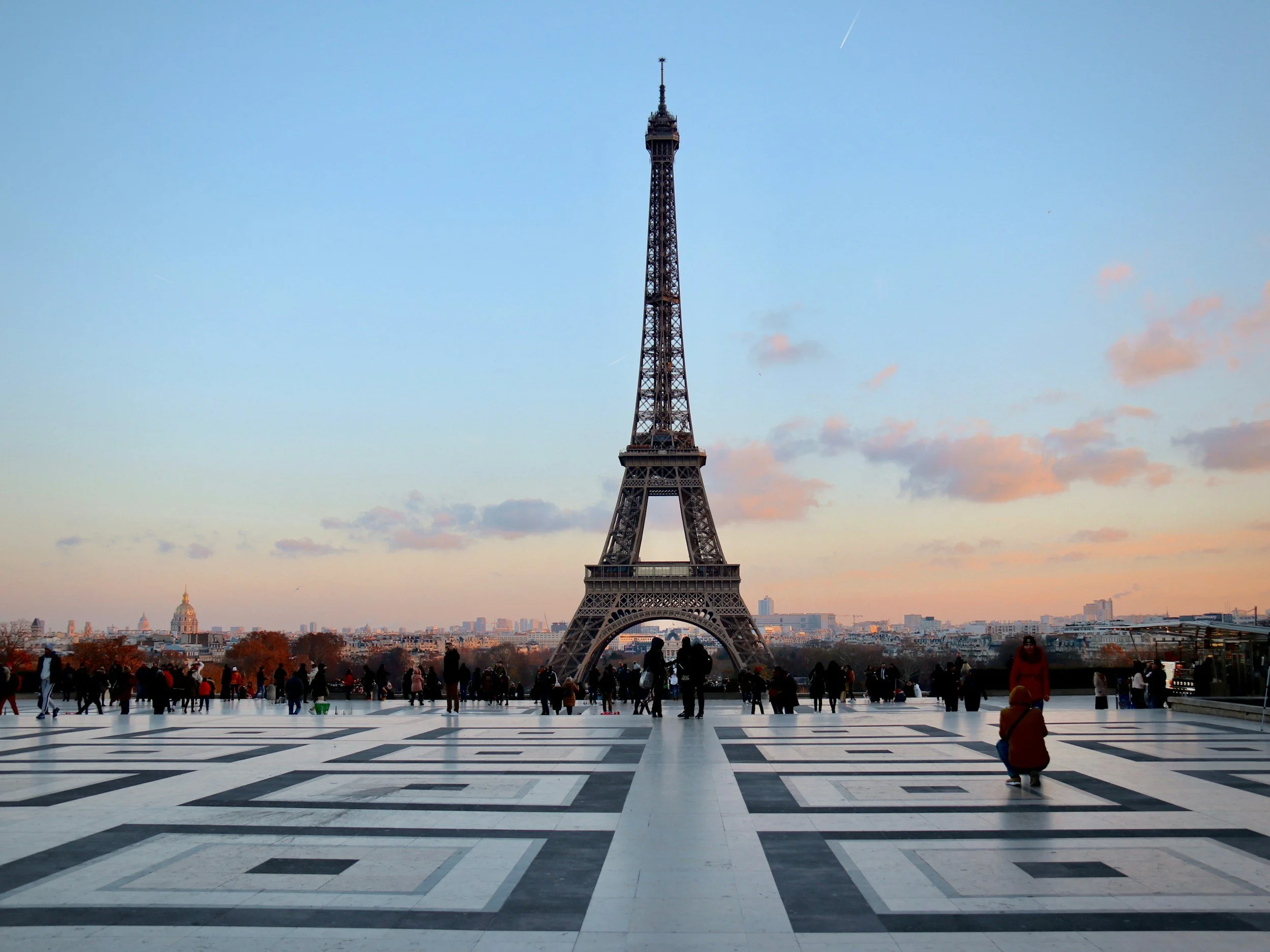 View of the Eiffel Tower in Paris, France, during sunset with a crowd of people gathered in the foreground on a large checkered plaza. The sky is clear with some clouds and a visible contrail.