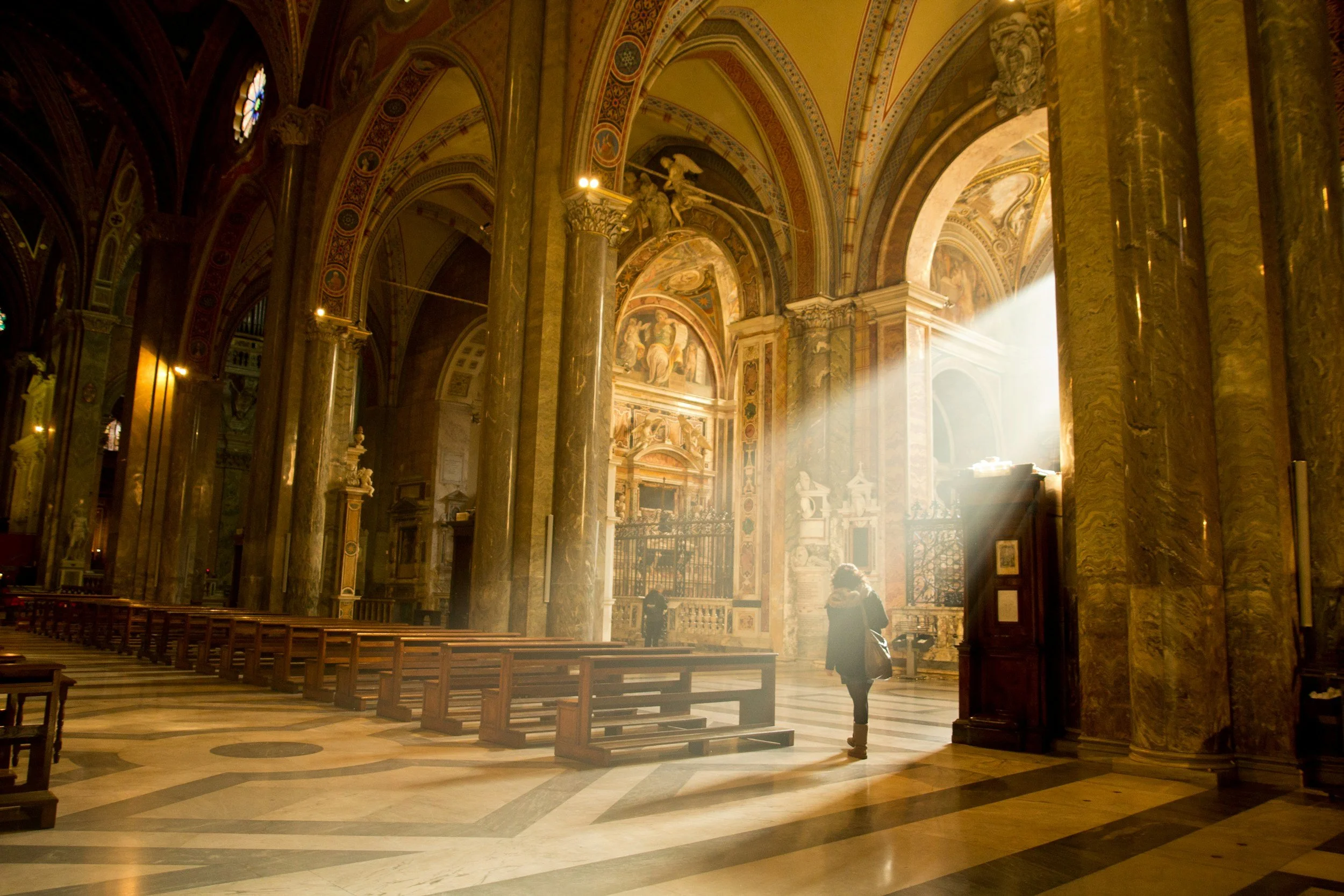 Interior of a grand church with tall marble columns, ornate painted ceilings, and sunlight streaming in through windows. A few people are present inside.