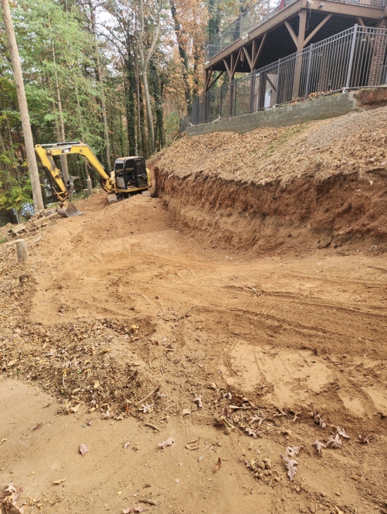 A construction site with a small yellow excavator digging soil along a dirt pathway surrounded by trees and a building with a deck and railing.