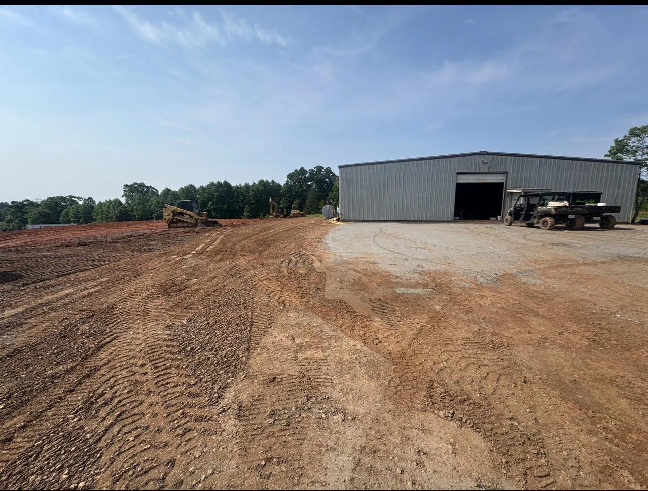 Construction site with a large metal warehouse building, construction vehicles, and a cleared dirt area.
