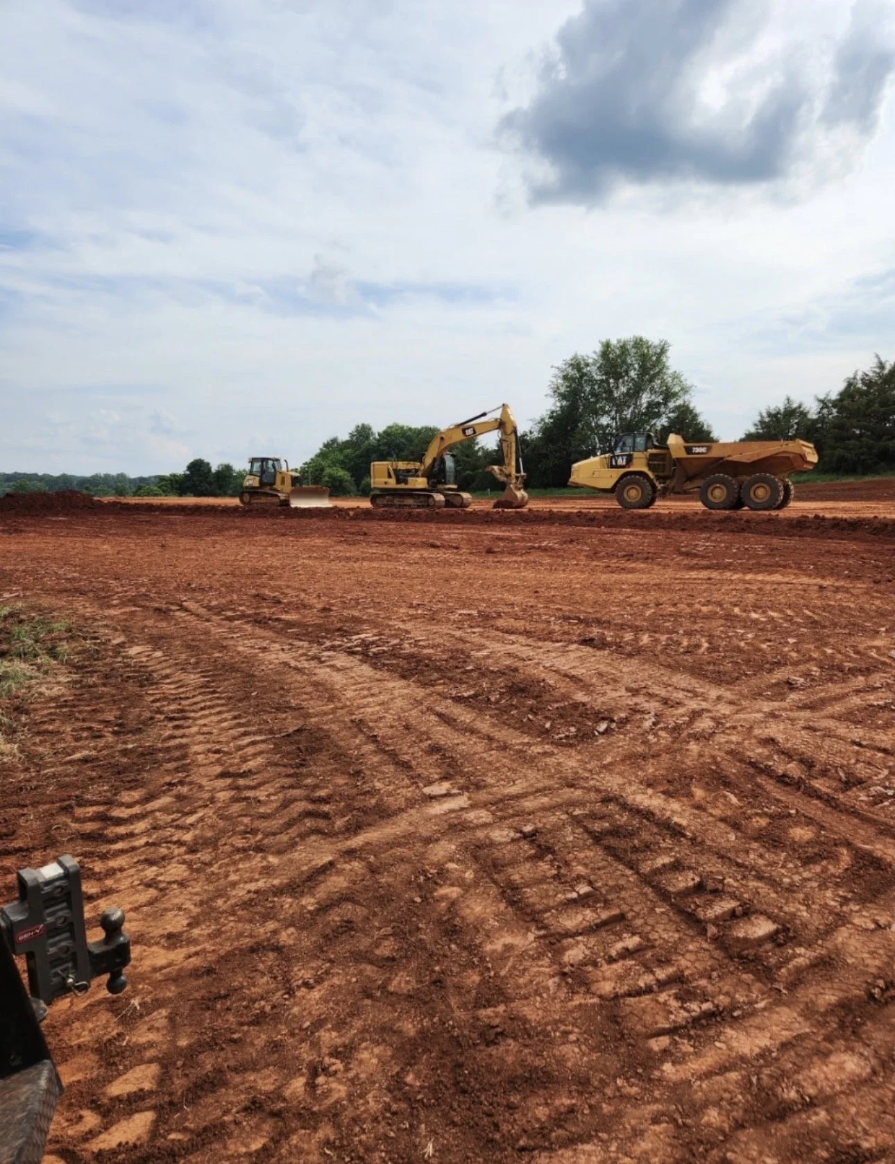 Construction site with three yellow heavy machinery vehicles on reddish-brown dirt ground, trees in the background, and a partly cloudy sky.