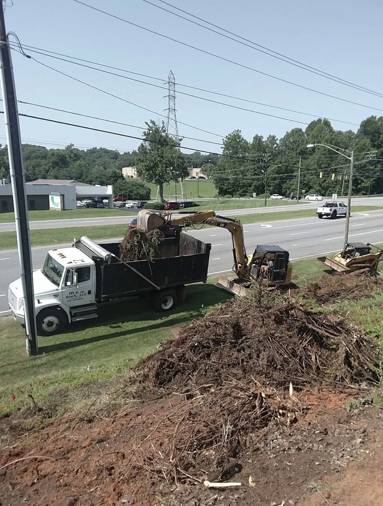 A construction vehicle with a hydraulic arm loading dirt and roots into a truck on a roadside, with trees, cars, power lines, and a building in the background.