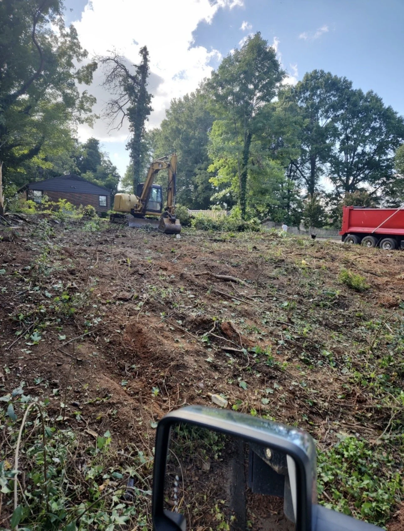A construction site with a yellow excavator bulldozing dirt and debris, surrounded by trees in a residential area with a house and a red dump truck in the background.