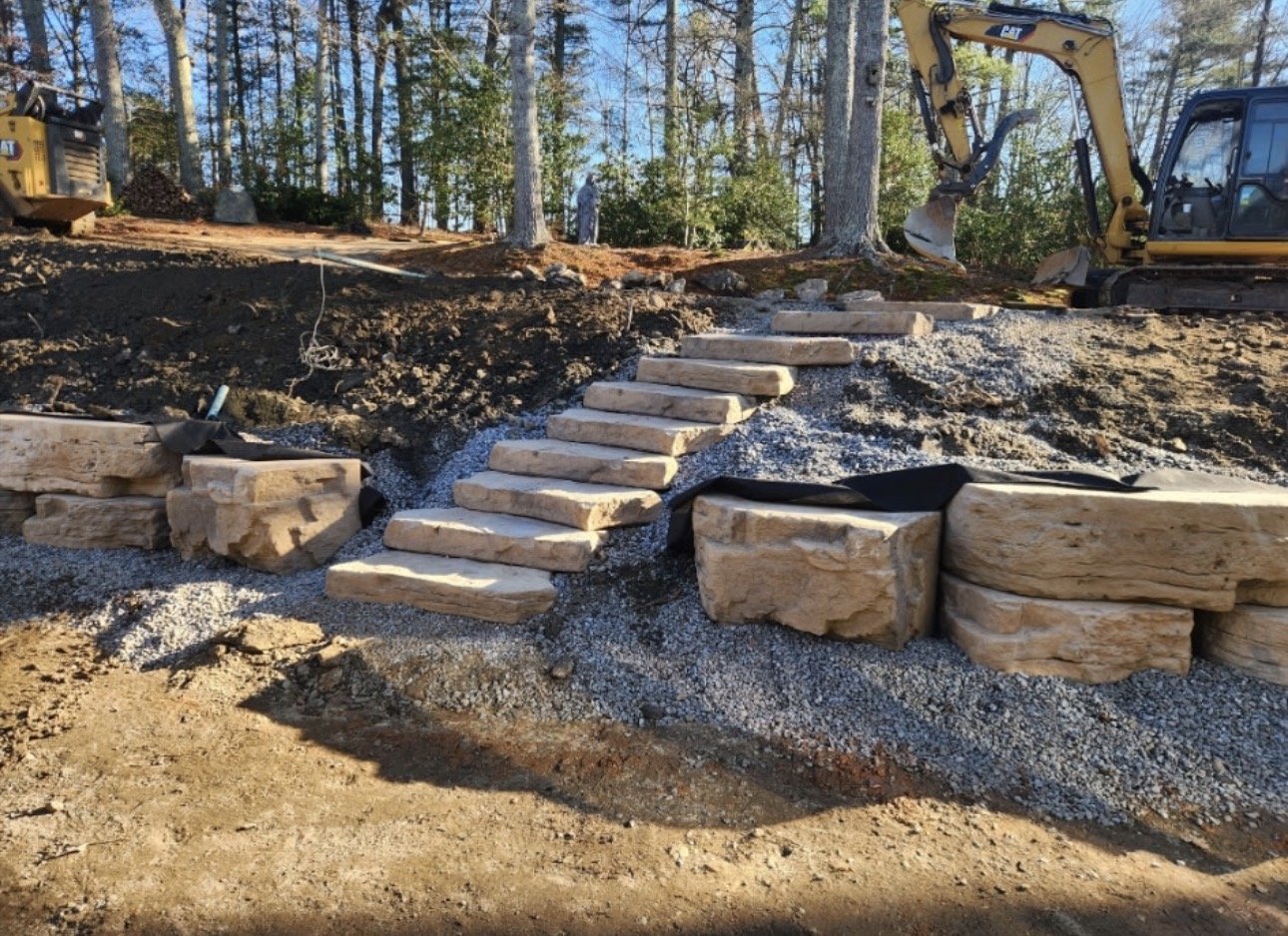 Construction site with stone steps and large rocks, two excavators, trees and dirt.