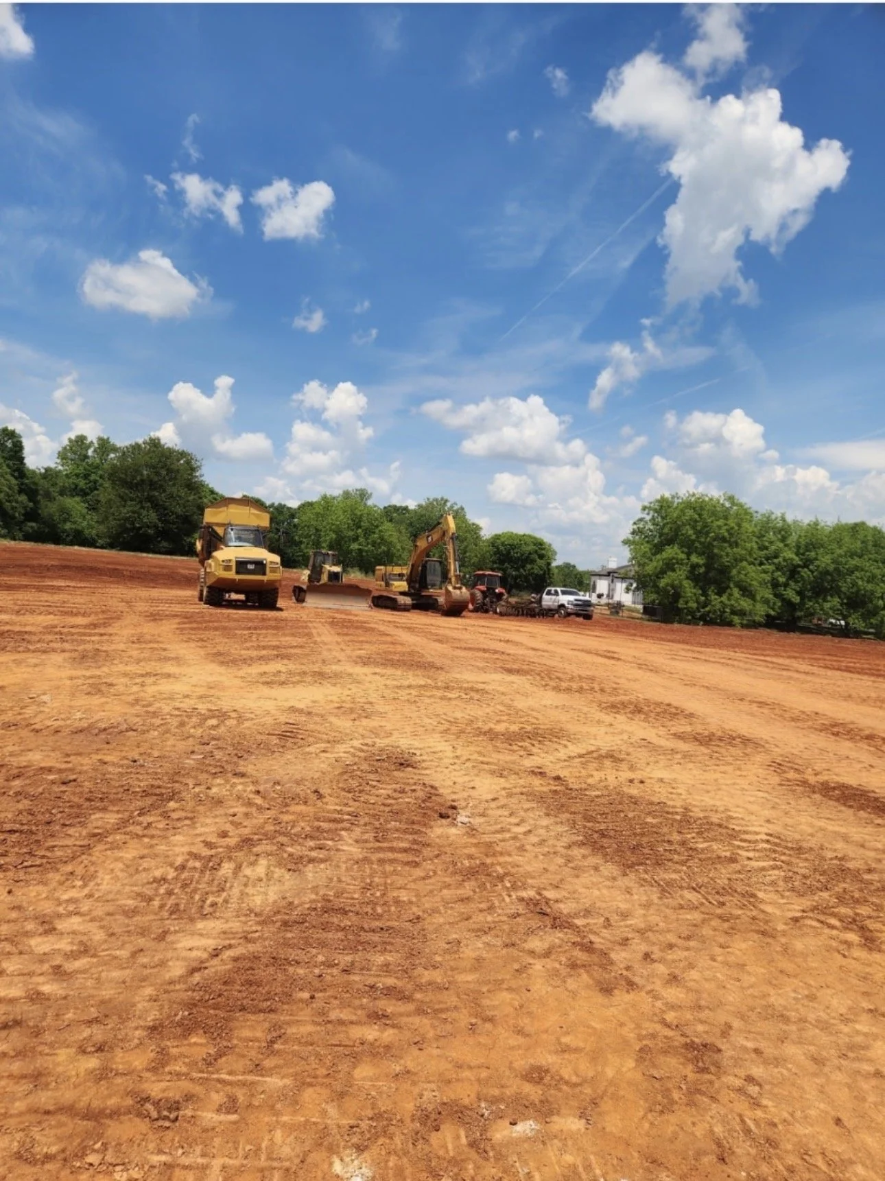 Construction site with yellow bulldozer, excavator, and tractor on red dirt under a blue sky with scattered clouds.
