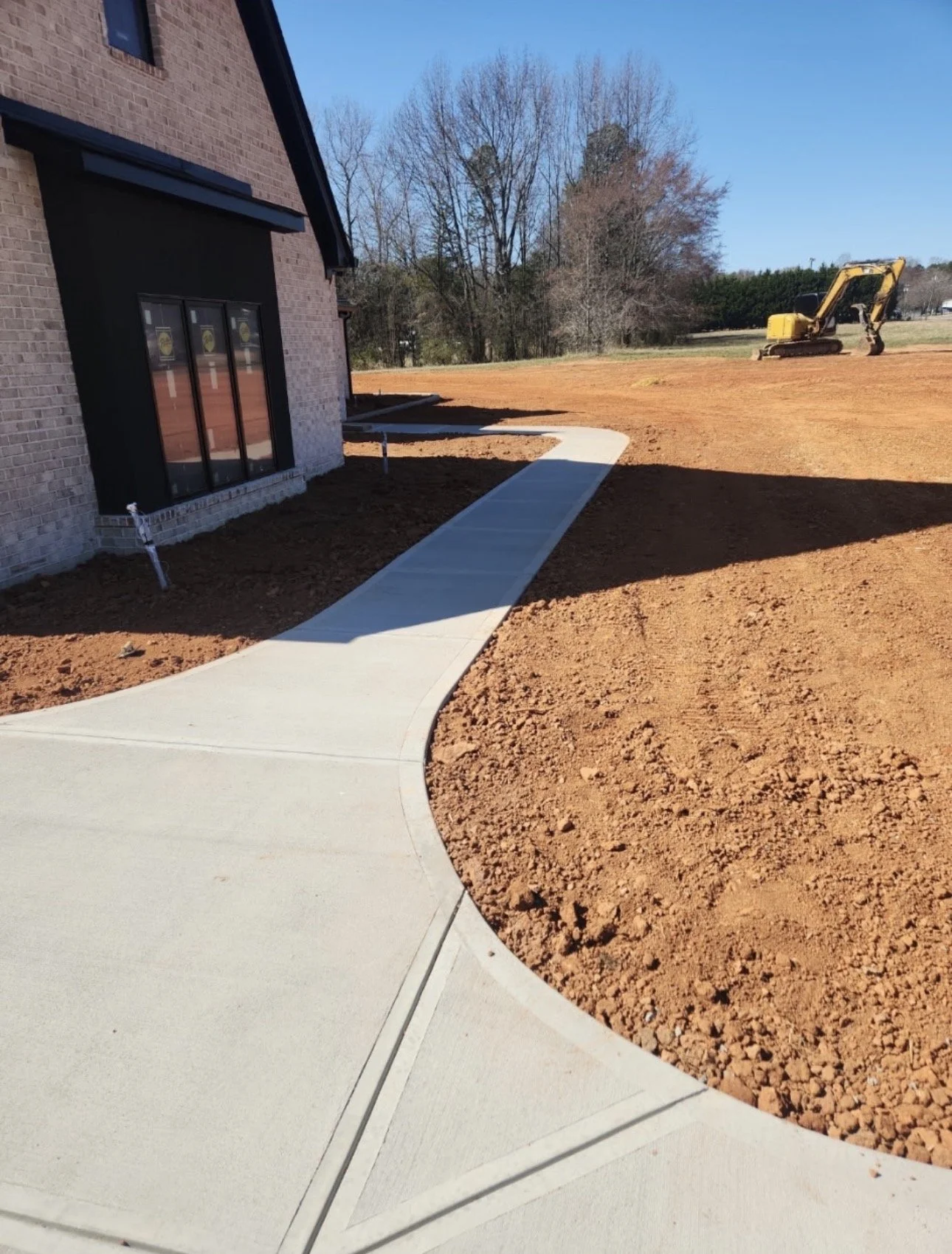 A concrete sidewalk curves around the side of a brick house, with bare trees and a yellow excavator in the background on a construction site. The ground is freshly tilled soil.