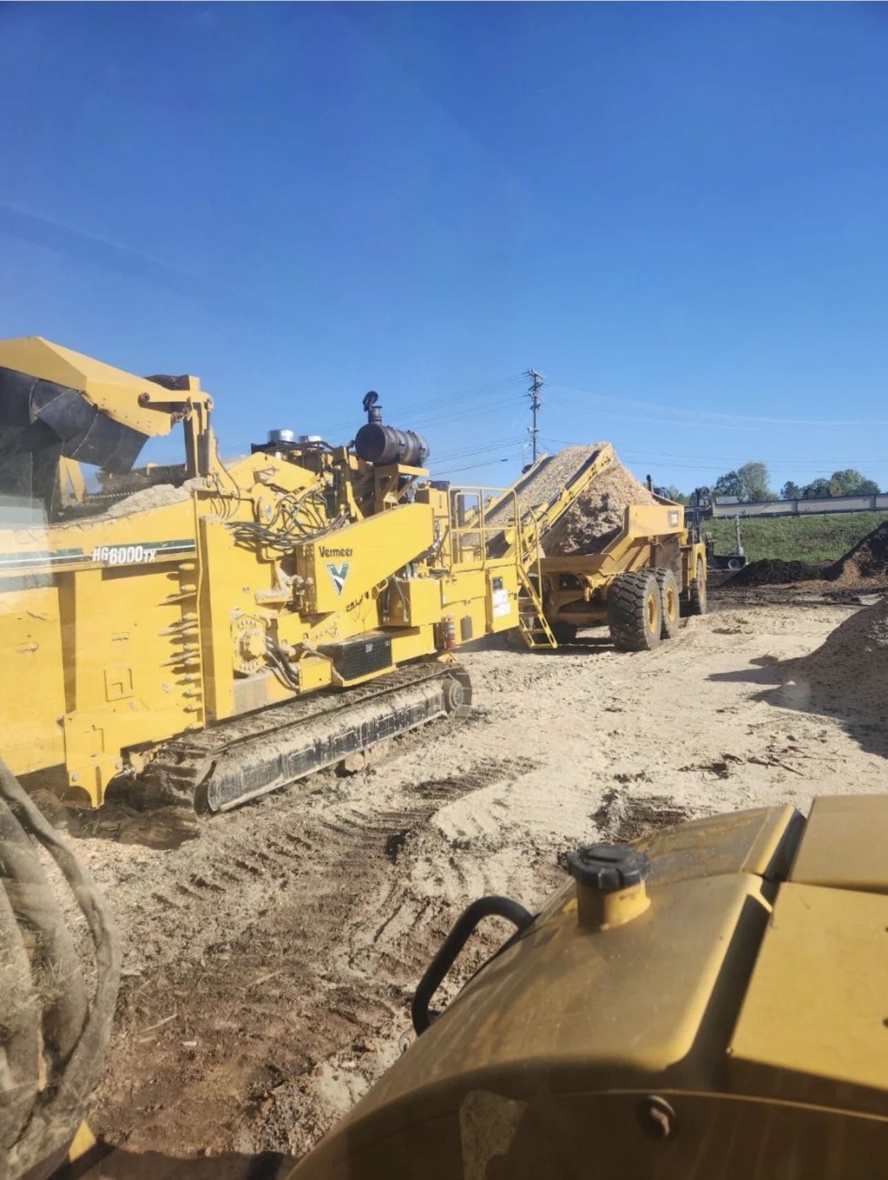 A construction site with yellow heavy machinery, including a road milling machine and a dump truck, on a dirt ground under a clear blue sky.