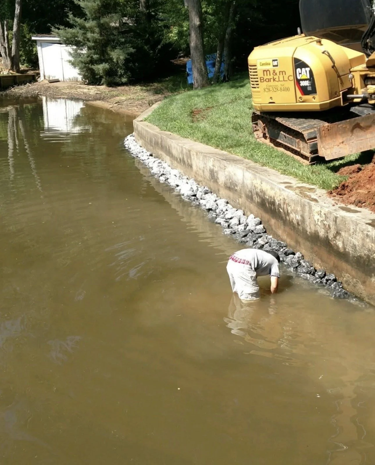 Person working by a water body, bending over near the shore, with construction equipment and trees in the background.