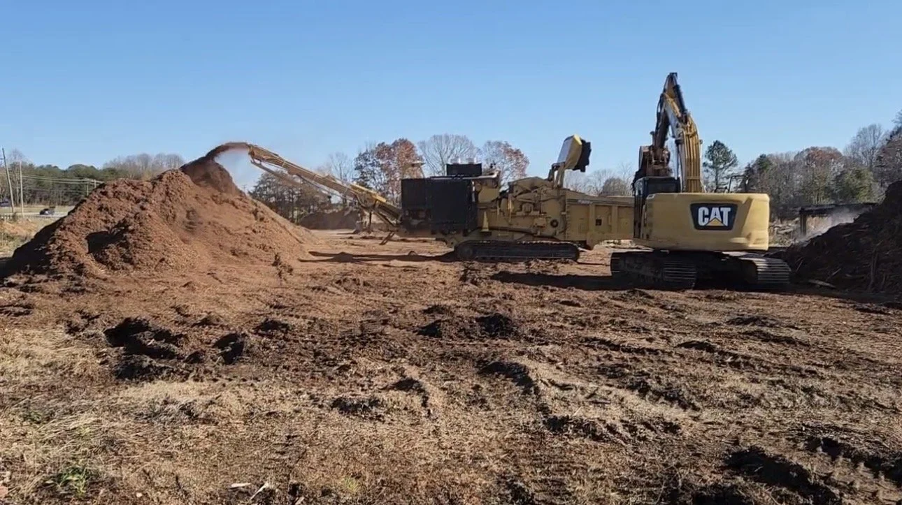 A construction site with large earth-moving machinery, including a CAT excavator, moving dirt and creating piles of soil against a backdrop of trees and a clear blue sky.