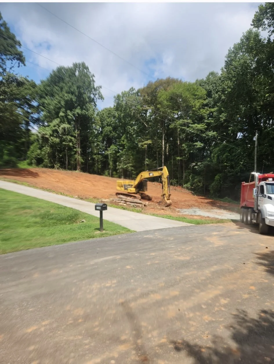 A construction site showing a yellow excavator on a dirt slope, with a dump truck parked nearby on a road. There is a sidewalk and a mailbox in the foreground, and trees with green foliage in the background under a partly cloudy sky.