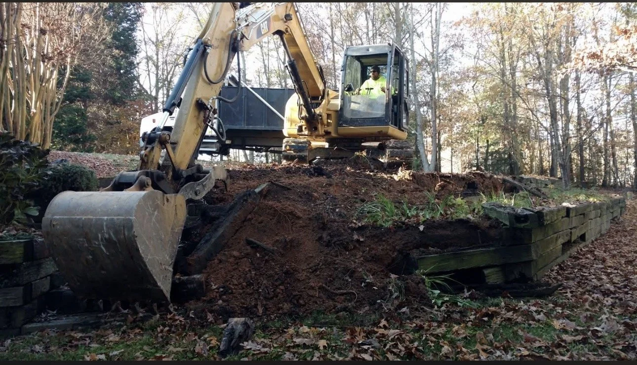 A person operating a small yellow excavator on a hill, surrounded by trees and fallen leaves, during daytime.
