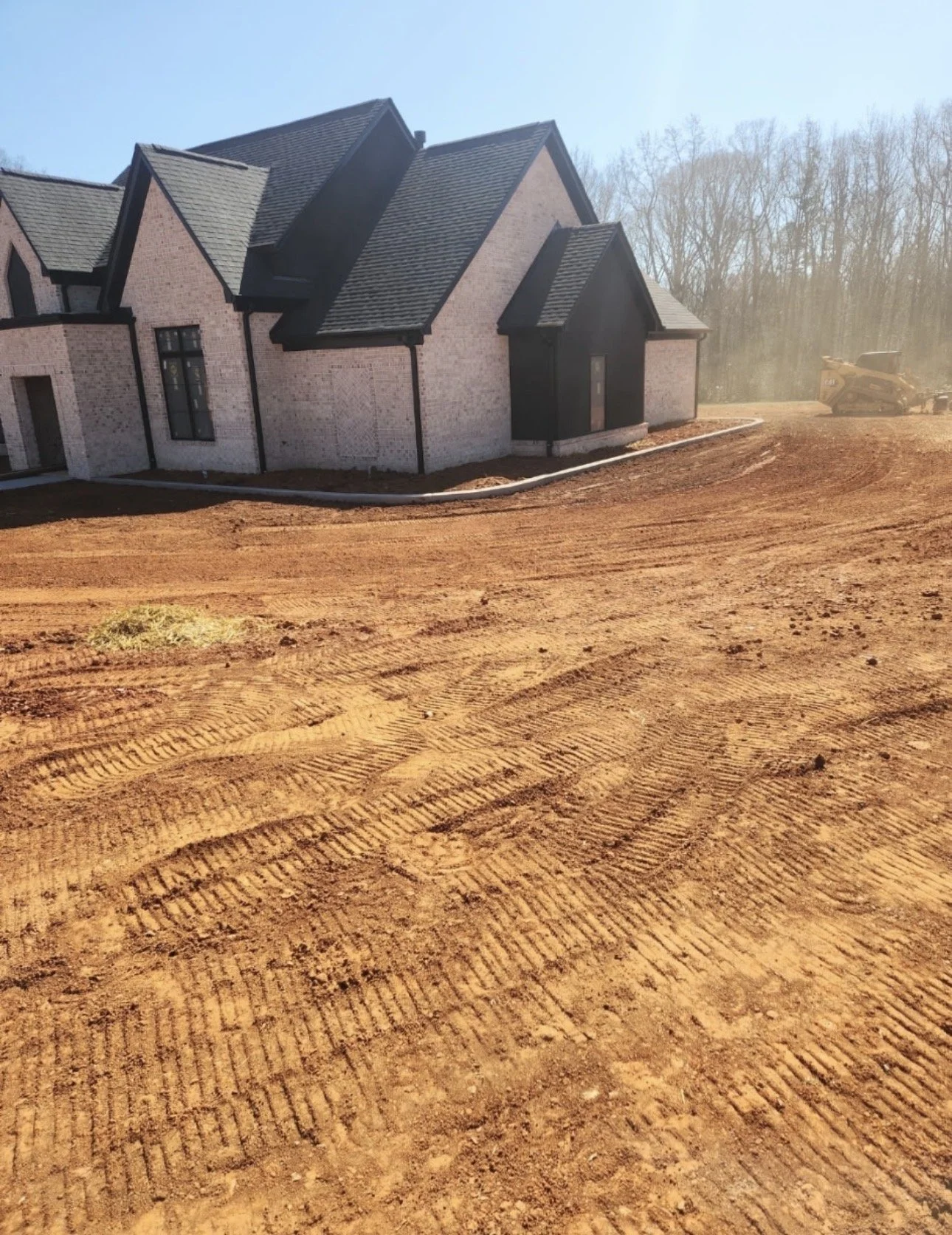 A newly constructed house with a brick exterior and black roof, with construction equipment working on the dirt driveway and clearing land in the background.