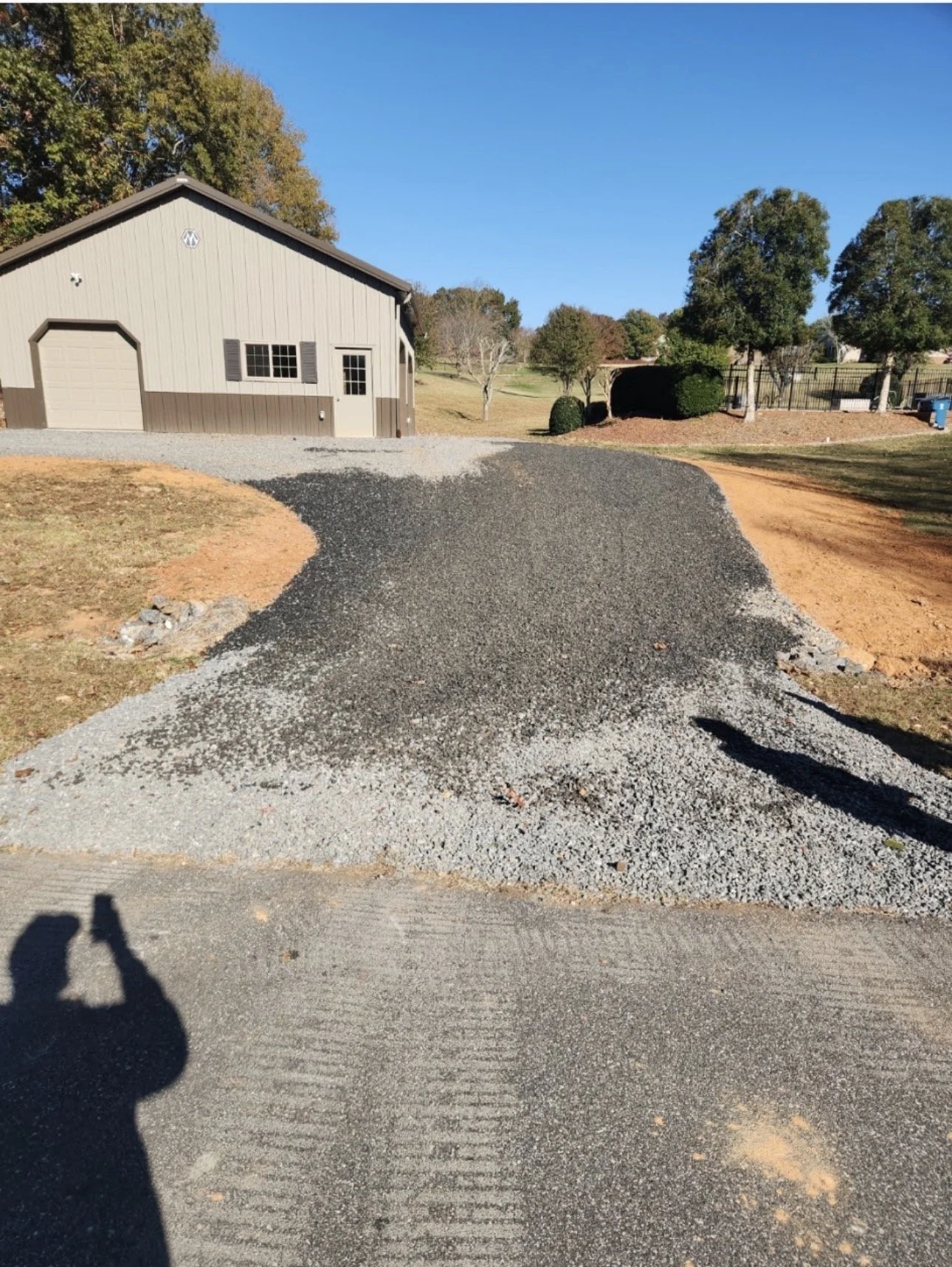 A gravel driveway curves towards a beige and brown shed with a closed garage door, set in a grassy yard with trees and a fence under a clear blue sky.