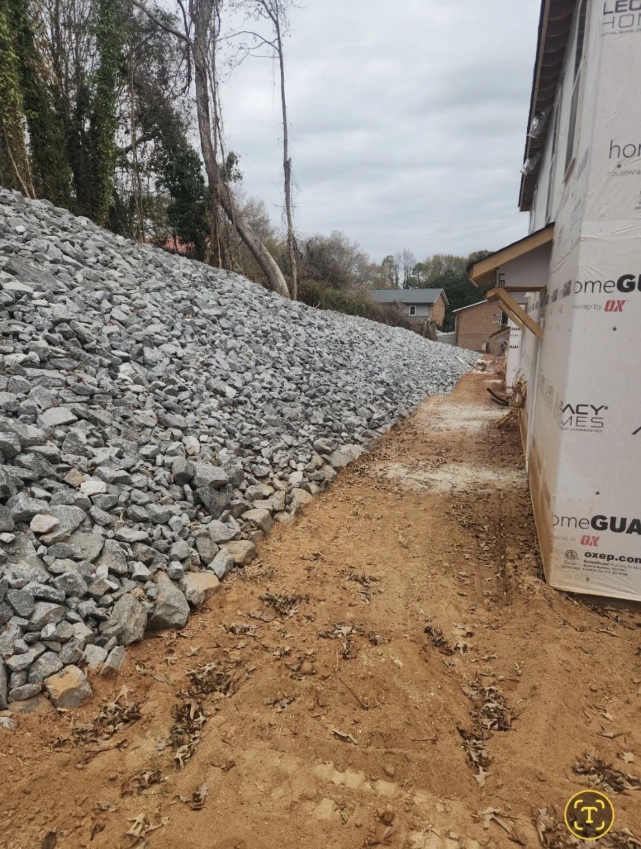 A construction site with a dirt pathway between a pile of gravel on the left and a building covered in weatherproofing on the right. The sky is overcast.