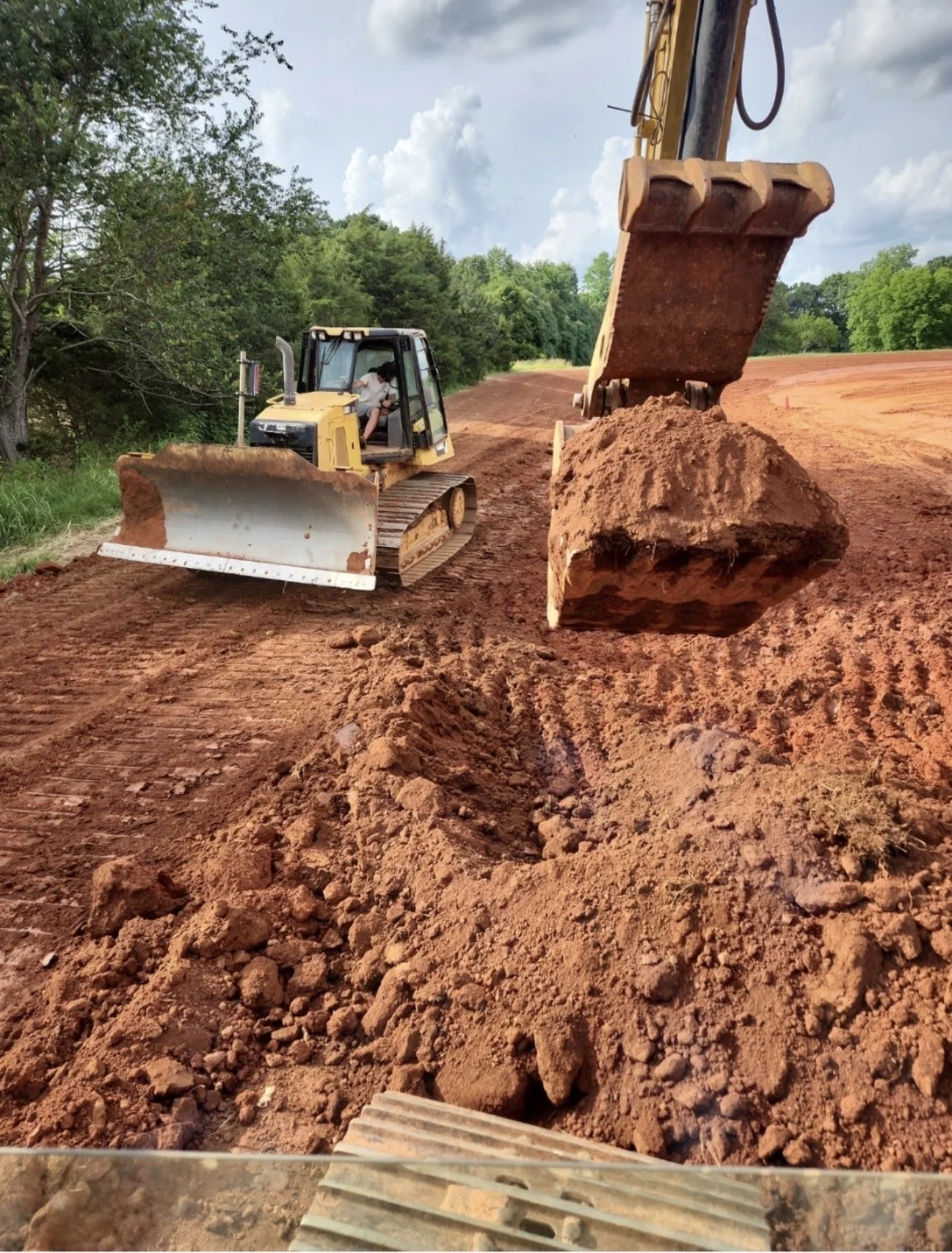 A bulldozer moving red soil on a construction site with trees and cloudy sky in the background.