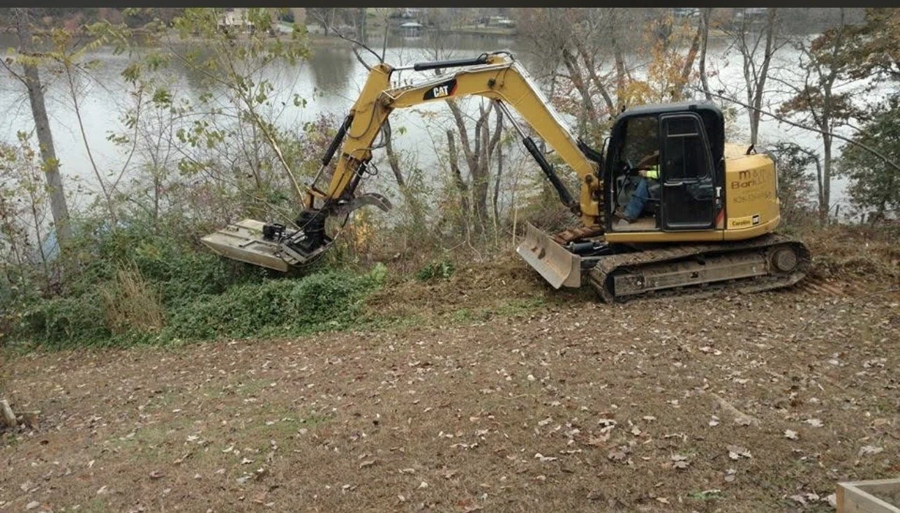 A small yellow Caterpillar excavator digging on a dirt surface near trees and a body of water.