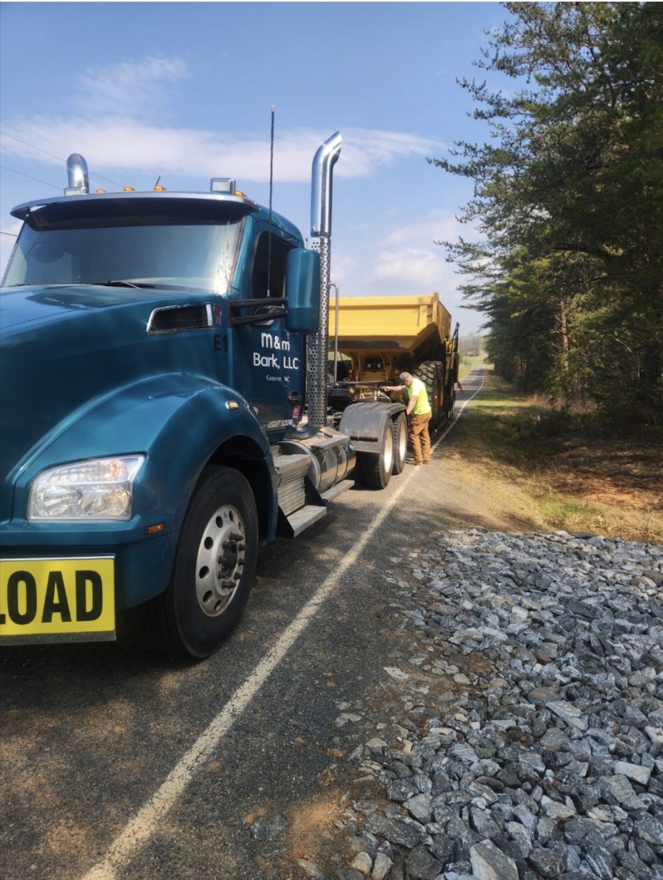 A large blue semi-truck with a yellow load, parked on the side of a rural road, with a man inspecting or working on the truck. The truck is parked near trees and a pile of rocks is in the foreground.