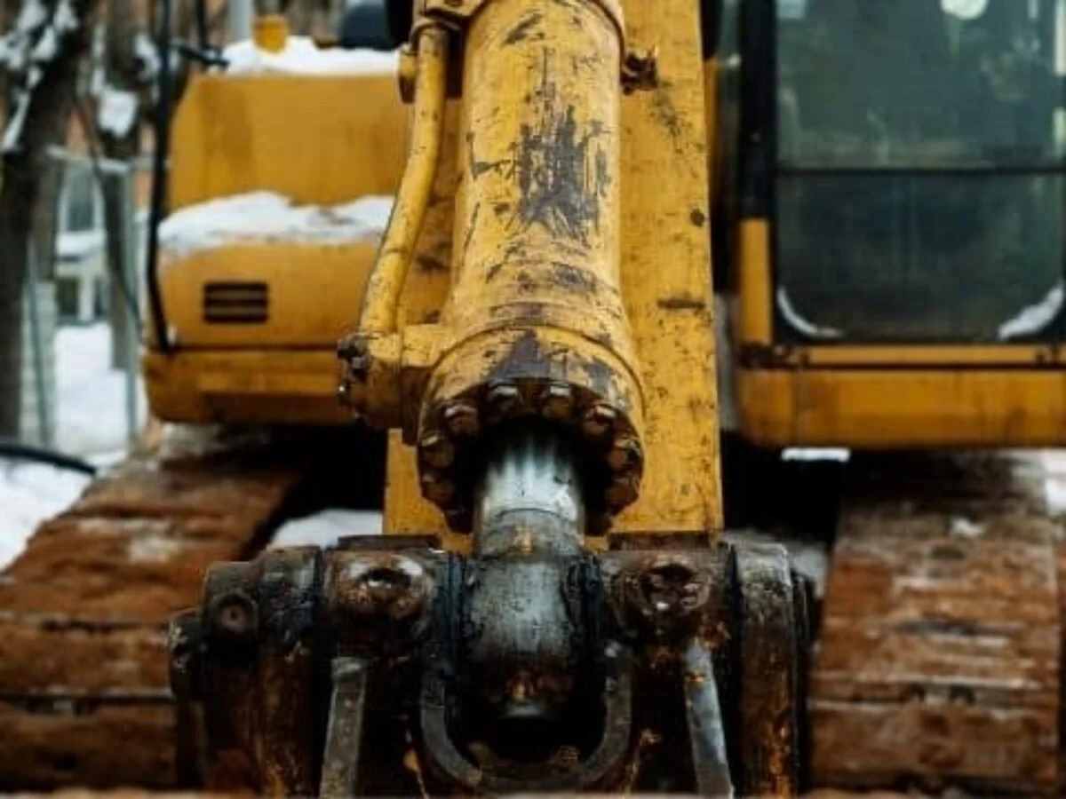 Close-up of the front arm and bucket of a yellow excavator with signs of rust and use, on a construction site with snow.