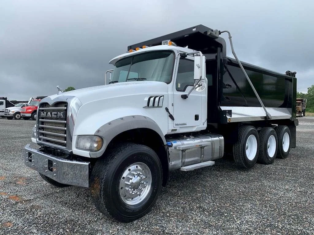 White dump truck with black bed parked on gravel lot under cloudy sky, multiple other vehicles in background.