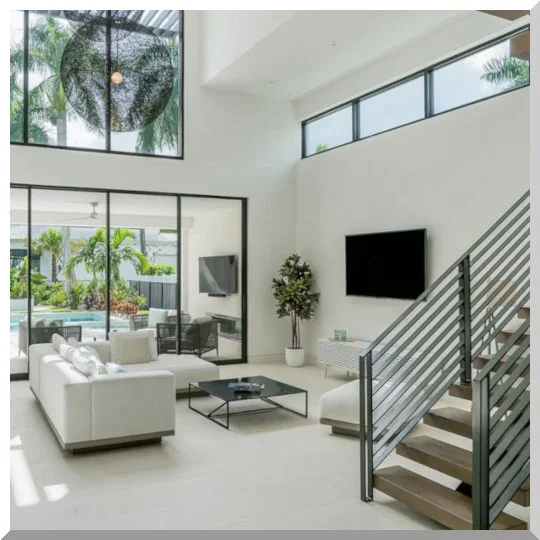 Modern living room with white furniture, a black coffee table, a wall-mounted flat-screen TV, large windows, and an outdoor pool area visible through glass doors.