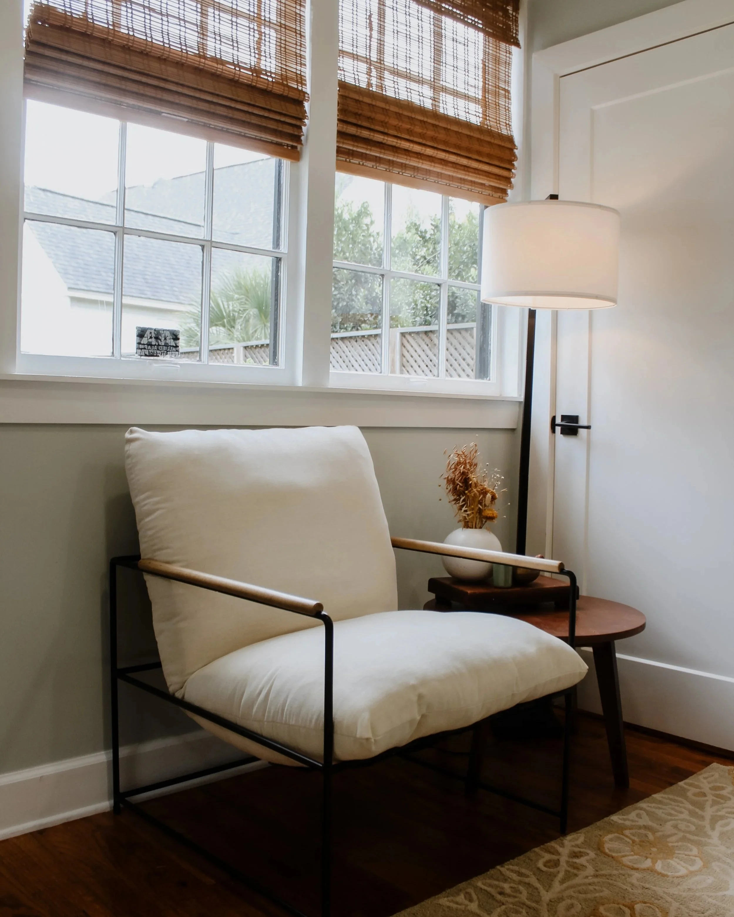 Modern bedroom corner with large window featuring bamboo blinds, black metal bed frame with cream-colored bedding, lighted white floor lamp, small wooden side table with decorative dried flowers, and a beige patterned rug.