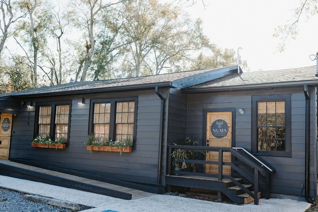 A dark gray building with multiple windows and a wooden door with a NUMA sign, surrounded by trees.