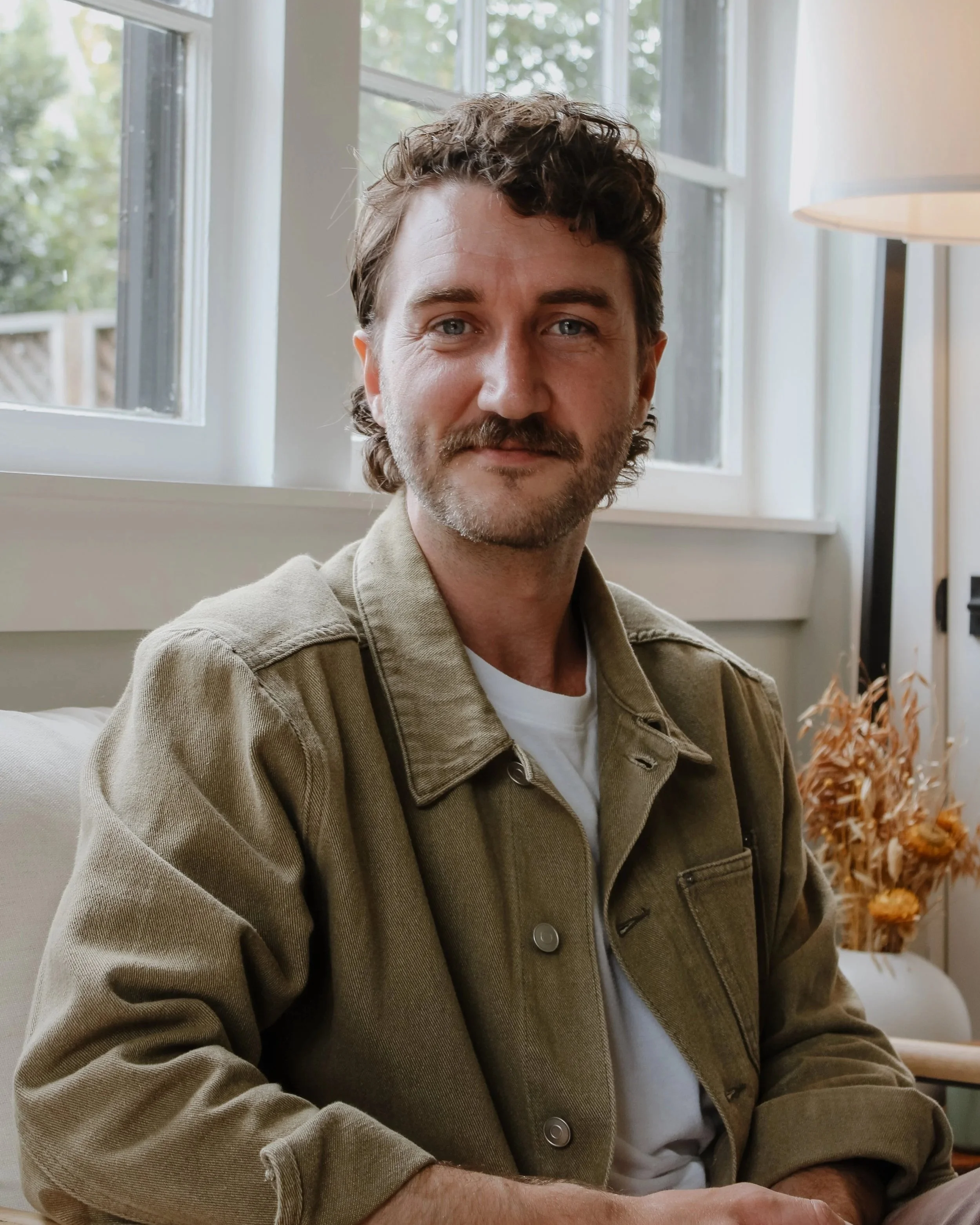 A man with brown curly hair, a beard, and blue eyes, sitting indoors near a window with a soft expression, wearing a beige jacket over a white shirt, with a lamp and dried flowers in the background.