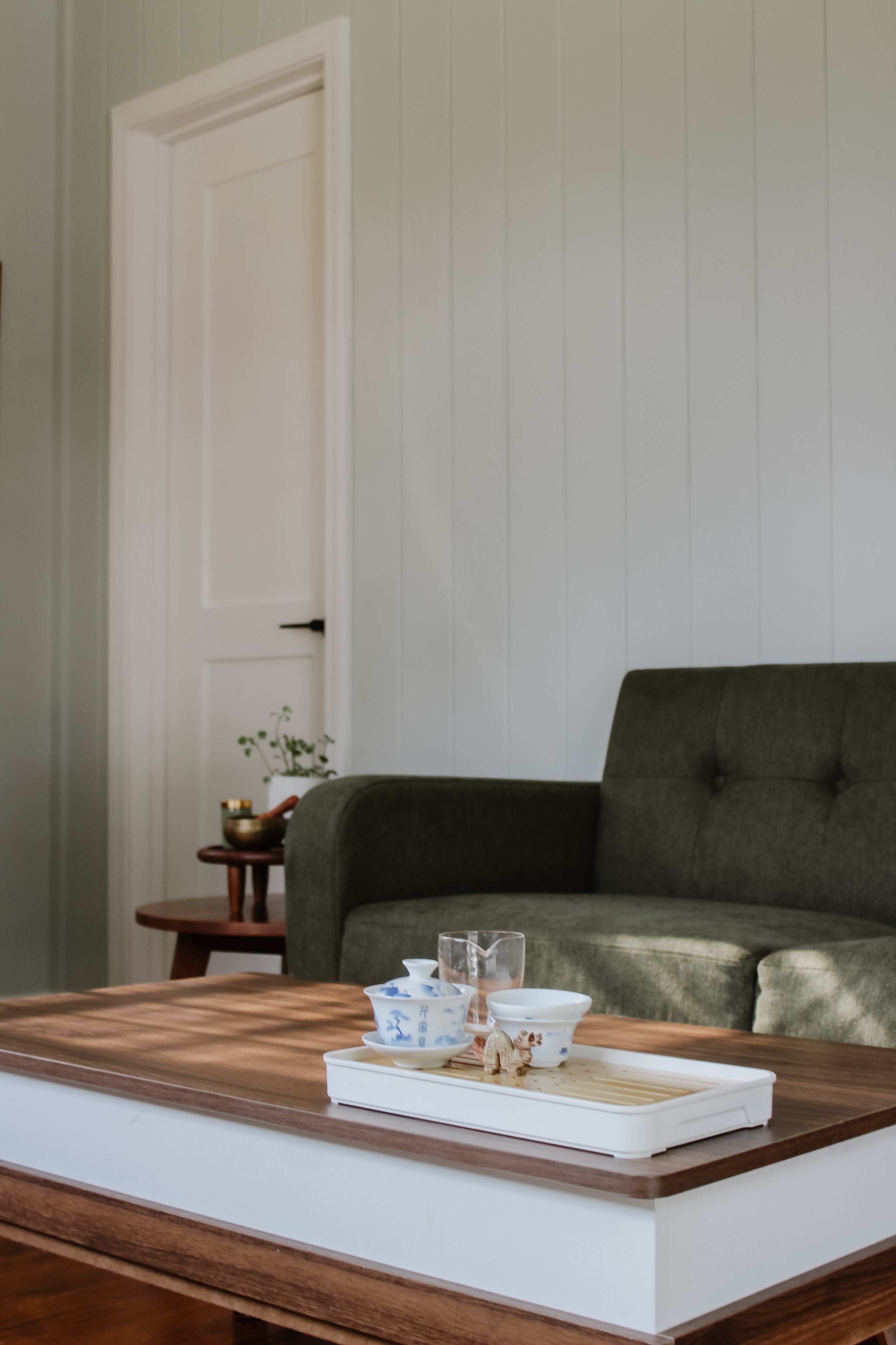 Office with sofa, wooden coffee table with tea cups, a small side table with a plant, and a closed white door against a light-colored wall.