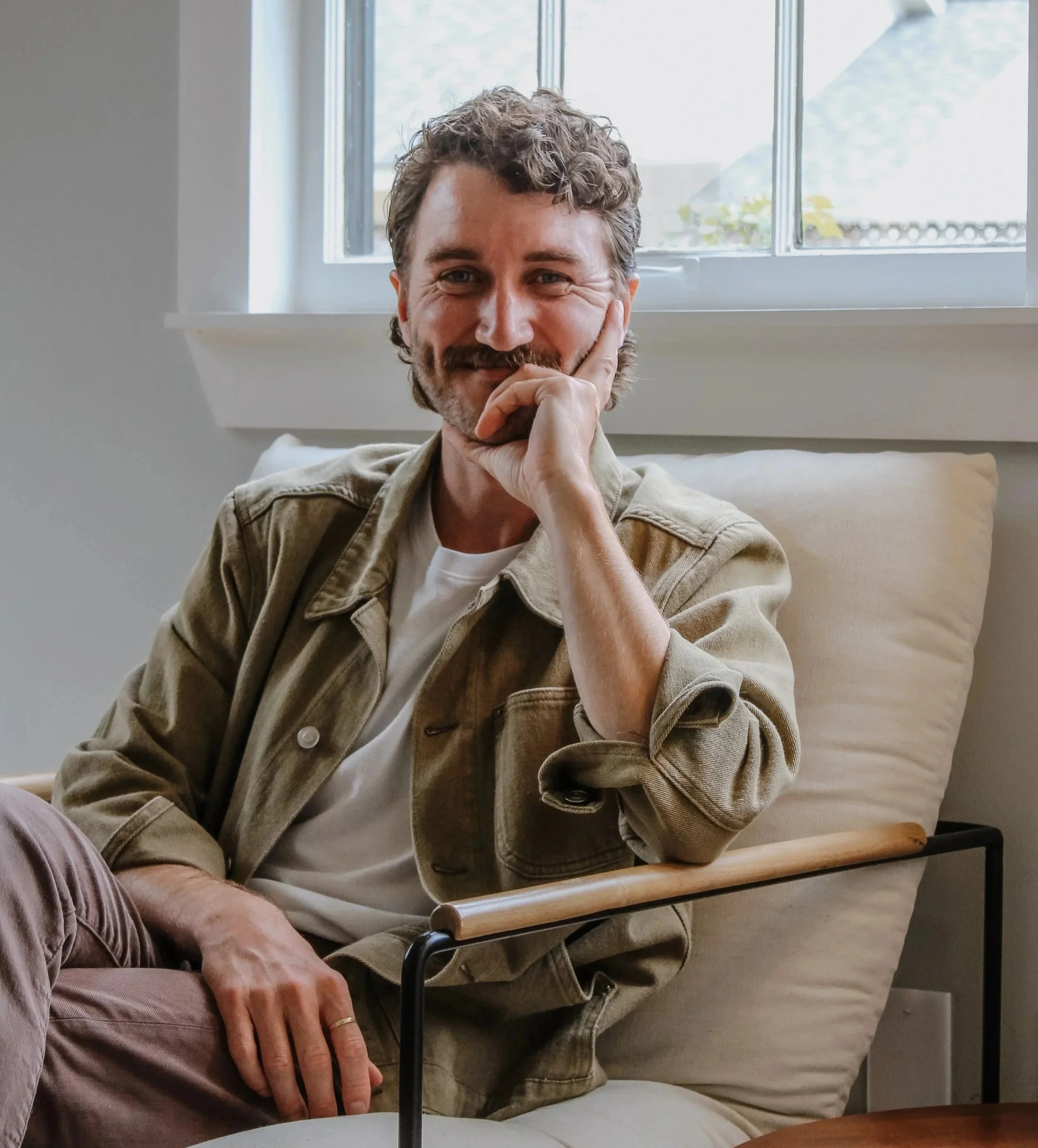 A middle-aged man with curly hair and a beard, smiling and resting his face on his hand, sitting next to a person with a light skin tone, wearing a beige jacket, outside near a window with a mountain and cloudy sky in the background.