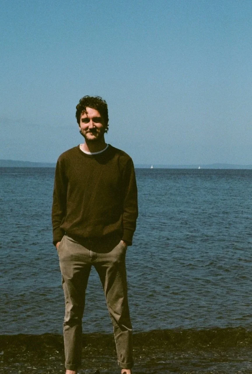 A young man with curly hair and a beard standing on a rocky shoreline by the water, smiling at the camera, with a body of water and sailboats in the background on a clear day.