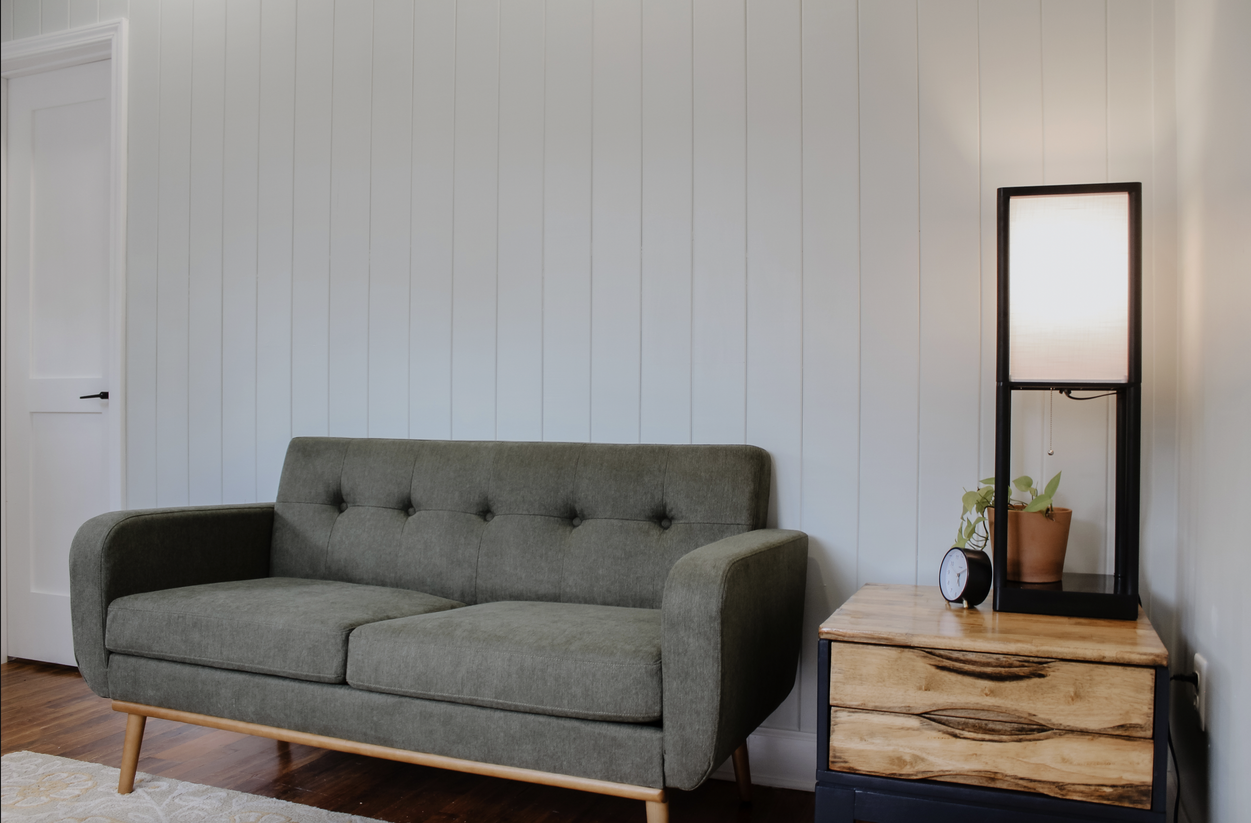 Living room with a gray upholstered sofa, a wooden side table with a potted plant, a black standing lamp, and a white paneled wall.