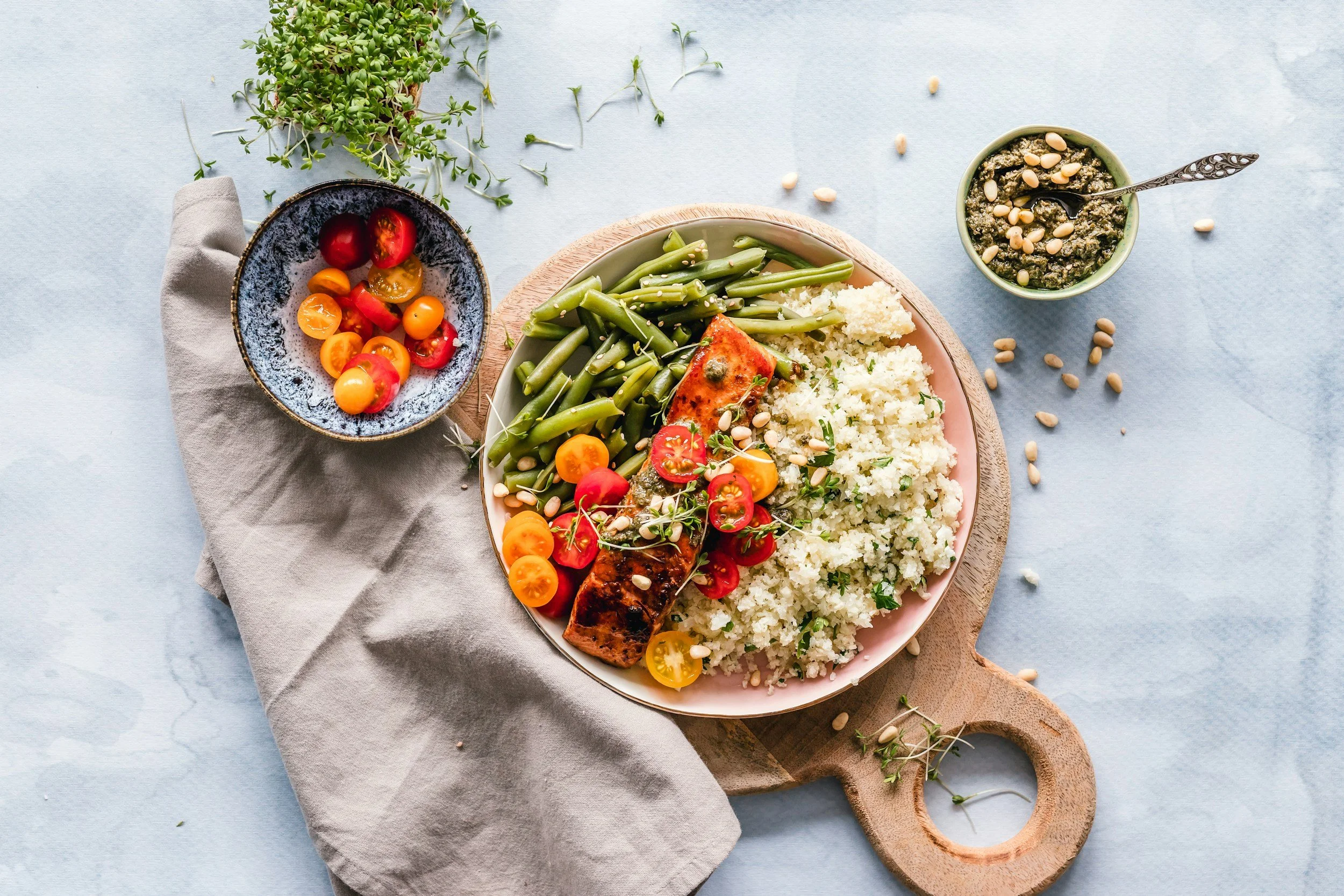 Plate of cooked salmon with cherry tomato garnish, green beans, and cauliflower rice, accompanied by a small bowl of pesto with pine nuts and a side of cherry tomatoes, on a wooden serving board and light blue tablecloth.