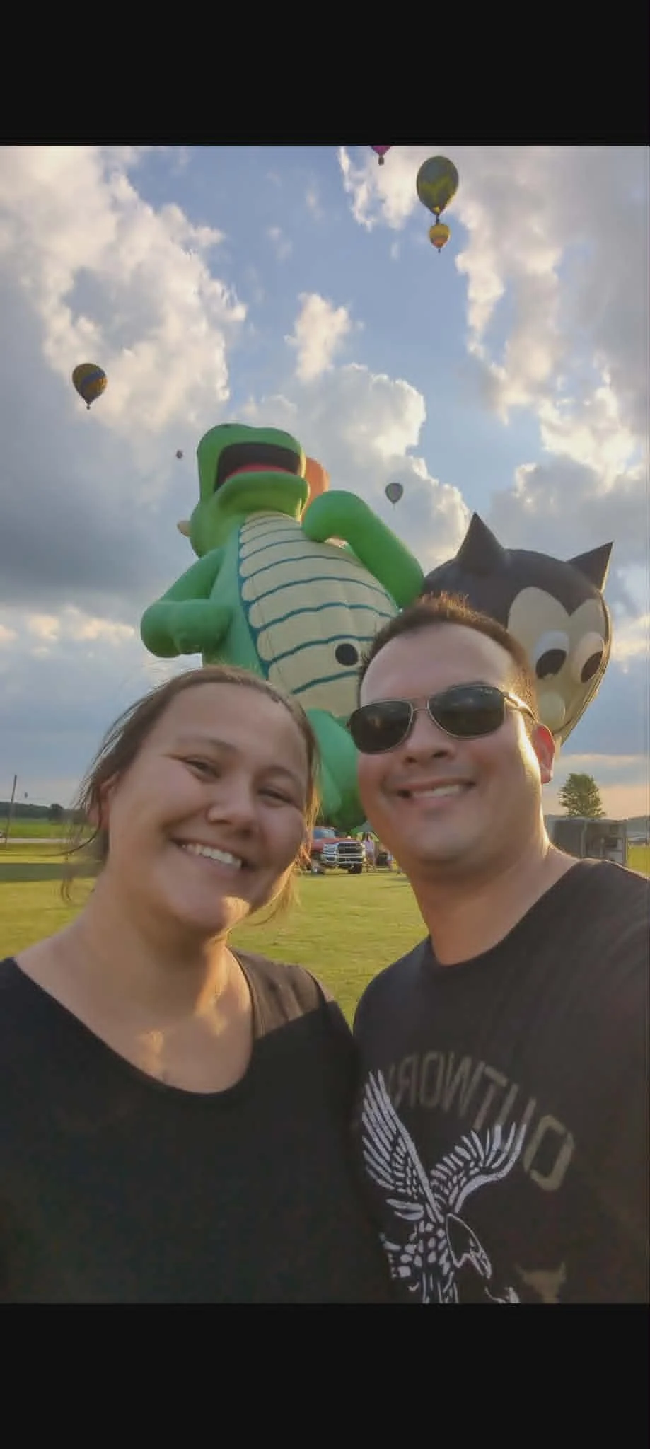 Two smiling people taking a selfie outdoors with hot air balloons and large animal-shaped inflatables in the sky behind them.