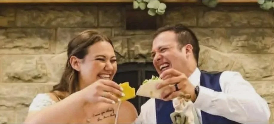 A man and woman laughing while sharing a taco in front of a stone fireplace.