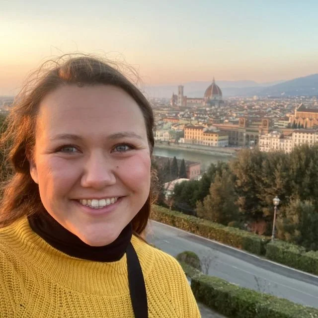 A young woman smiling outdoors at sunset with Florence, Italy, skyline and the Florence Cathedral in the background.