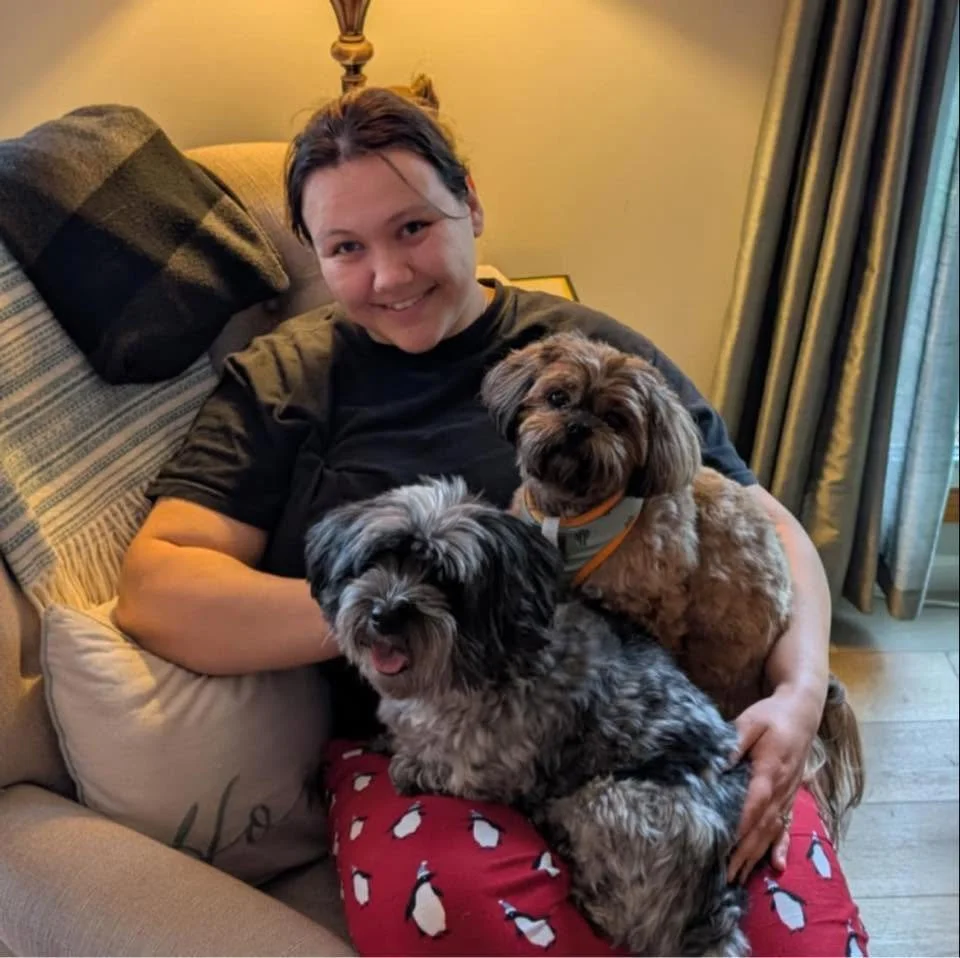 A woman sitting on a sofa holding two small dogs, one black and gray with a happy expression and one brown with lighter patches, in a cozy room with curtains and a lamp.