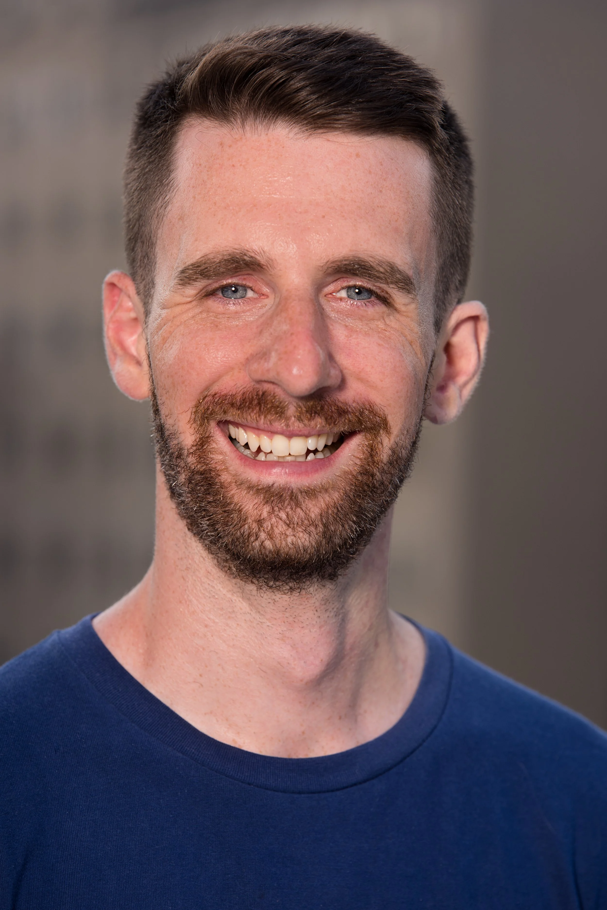 Close-up of a smiling man with short brown hair, light skin, blue eyes, and a beard, wearing a navy blue shirt, against a blurred gray background.