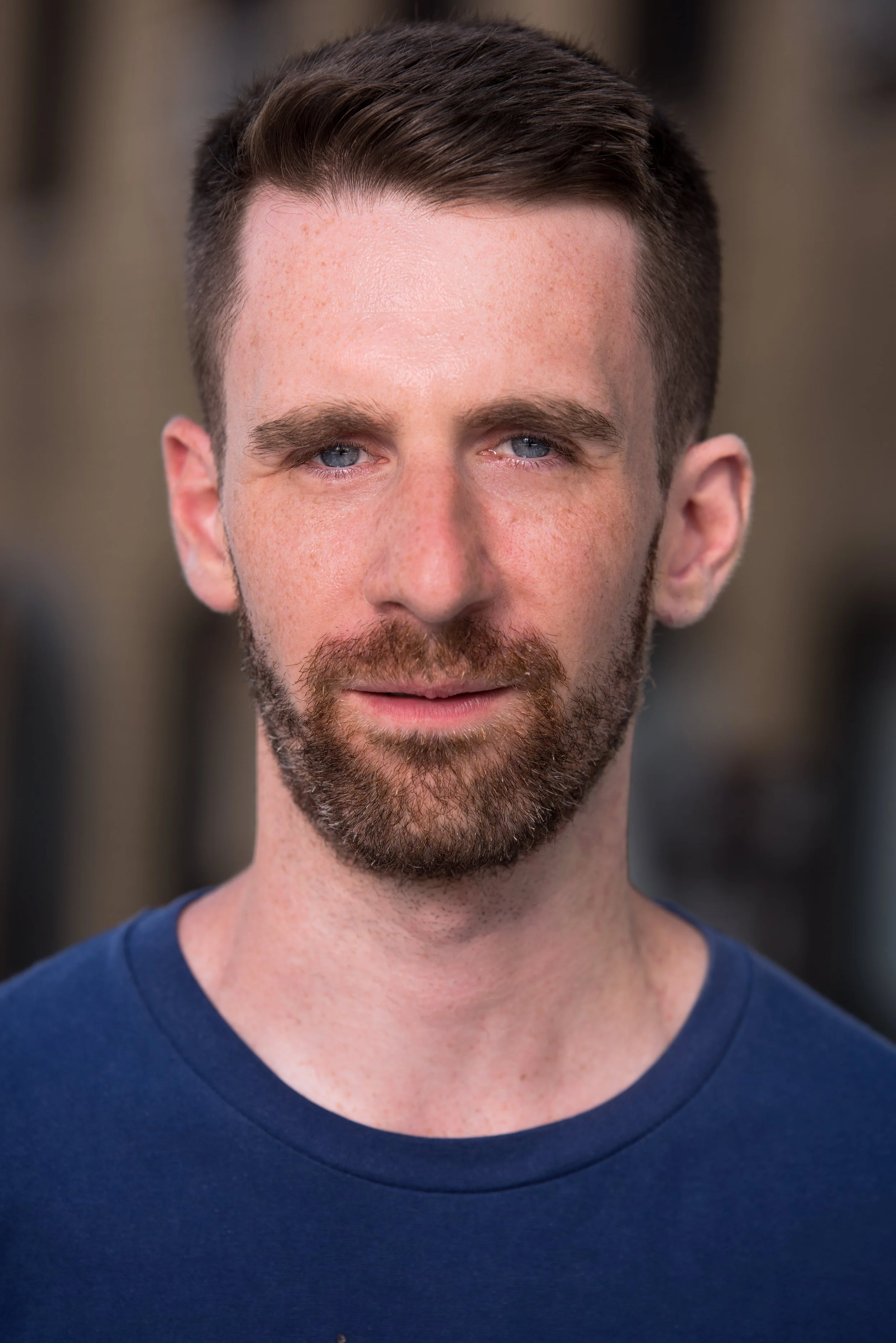 Close-up of a young man with fair skin, blue eyes, brown hair, and a beard, wearing a blue shirt, looking directly at the camera with a neutral expression.