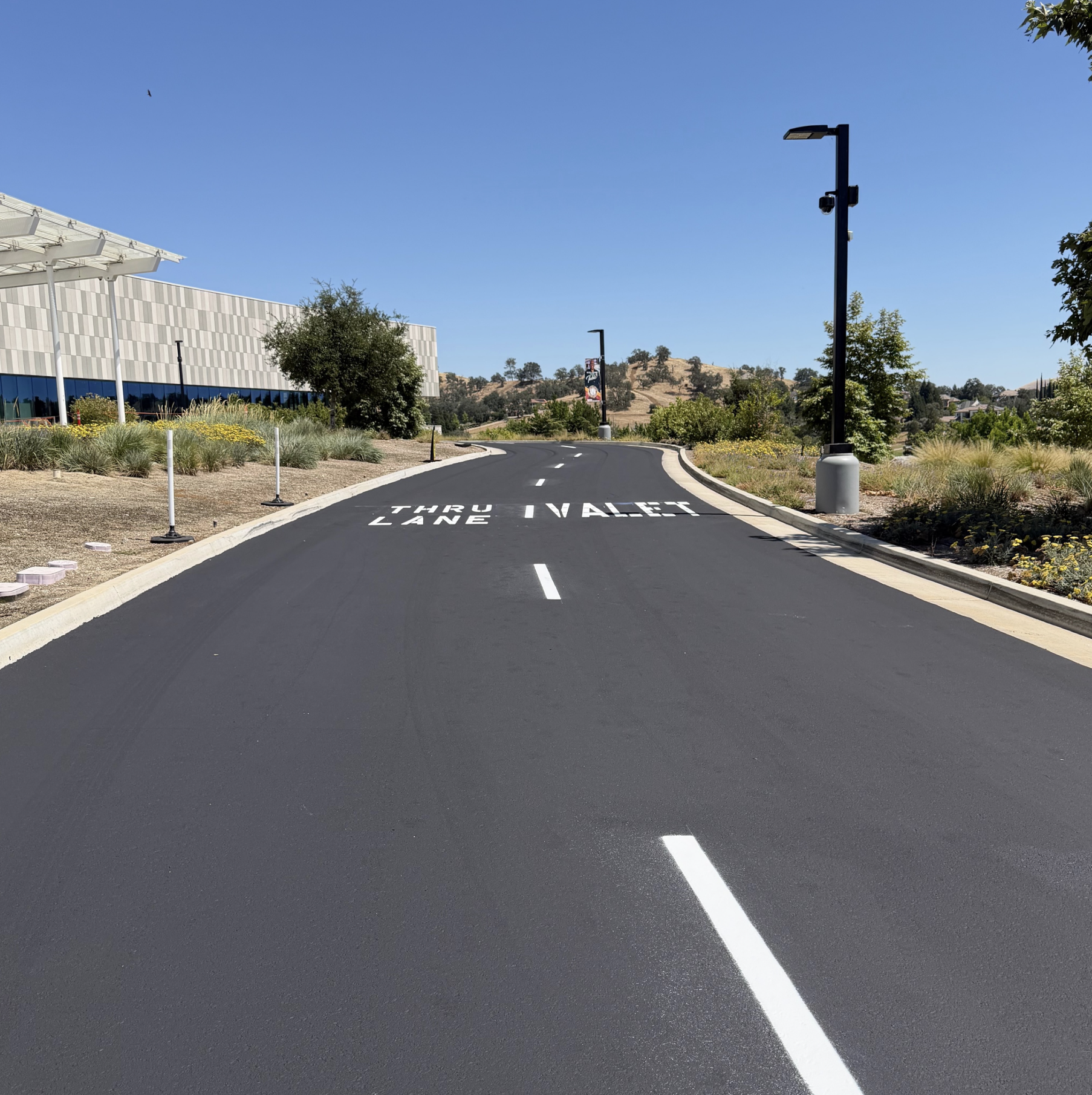 A paved road with white lane markings and signage that reads 'THRU LANES' on the ground, surrounded by landscape with trees, shrubs, and a modern building on the left, under a clear blue sky.