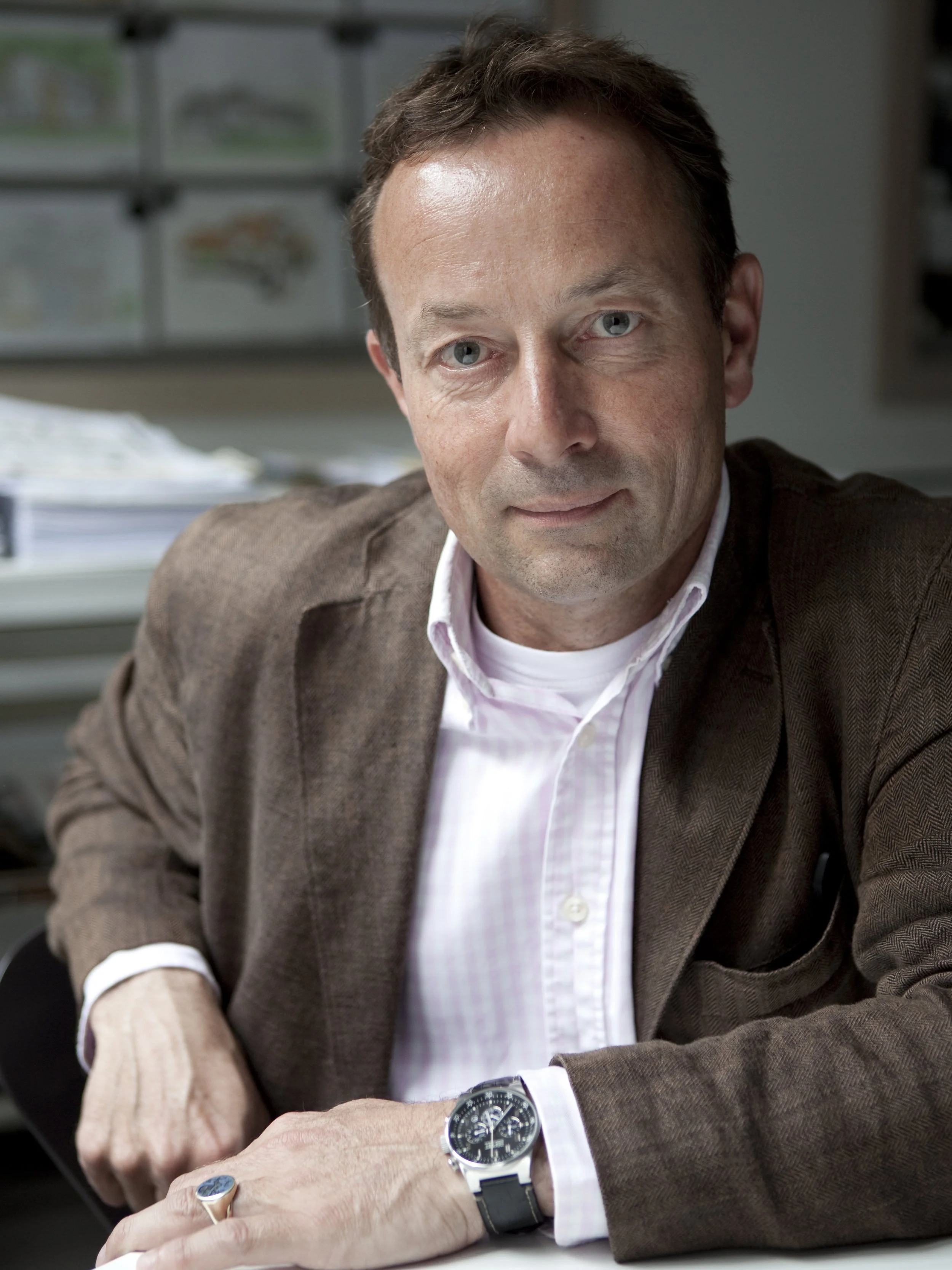 Man met blauwe ogen, bruin haar, witte overhemd en bruin colbert zit aan een bureau, draagt een horloge en een ring.